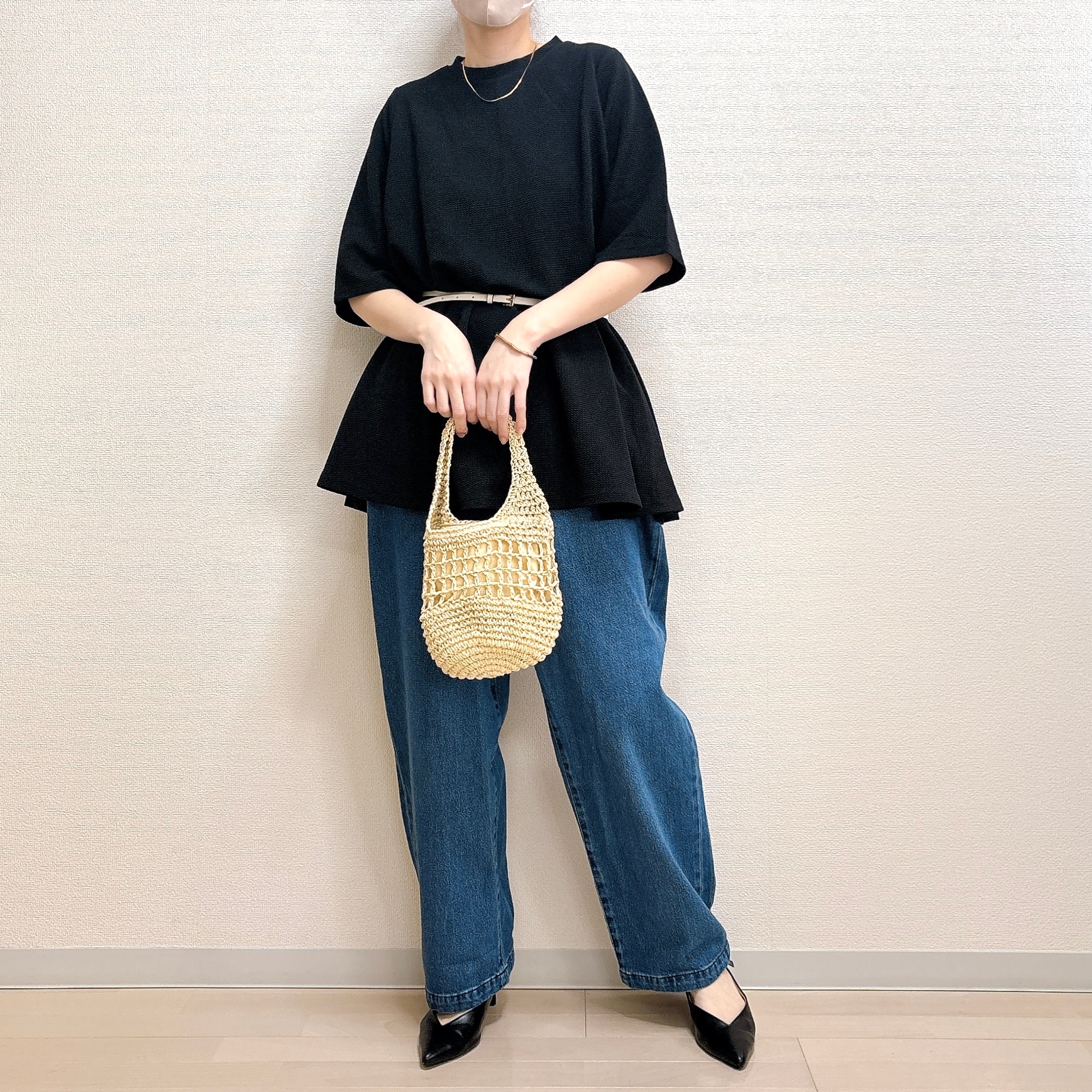 ゆったりした黒のトップスにデニムパンツ、ベージュの編みバッグを持った人が写っています。シンプルでカジュアルなスタイルです。