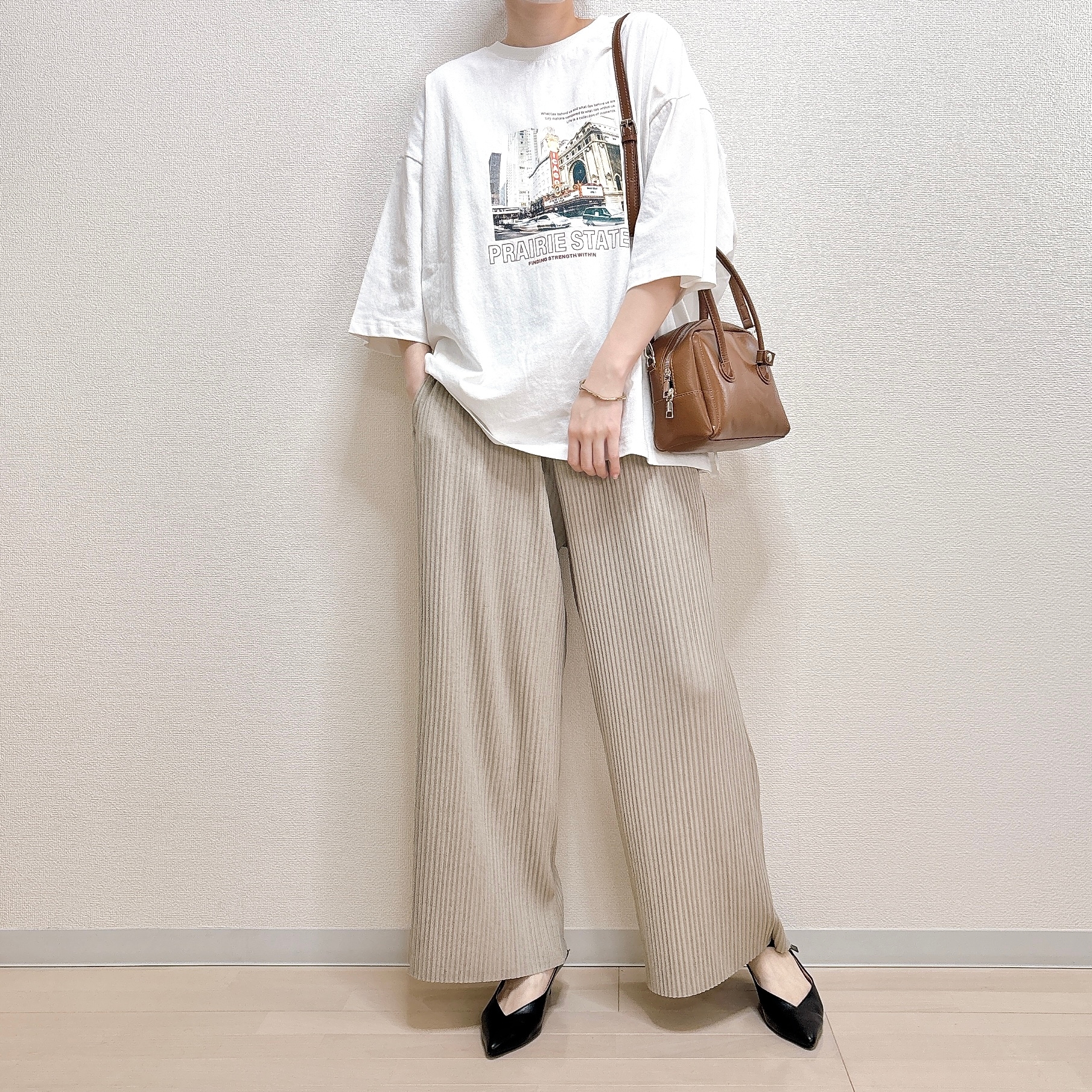 女性が白いプリントTシャツとベージュのワイドパンツを着用し、肩に茶色のバッグをかけて立っている。モダンカジュアルなスタイル。