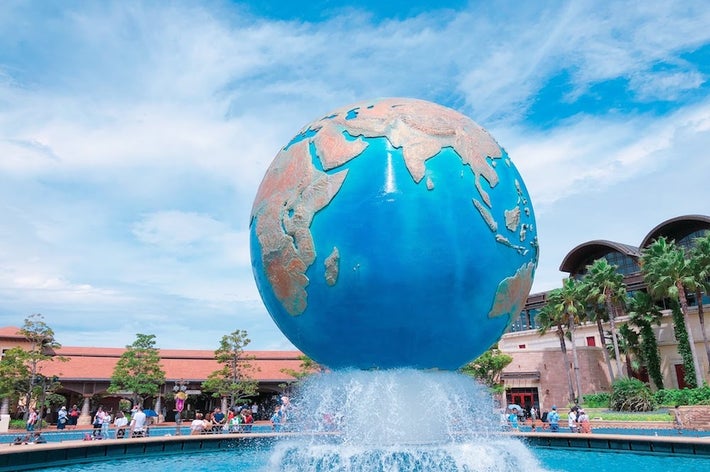 地球の形をした大きなオブジェが噴水の上にあり、周囲に人々が集まっています。背景には建物と木々があります。