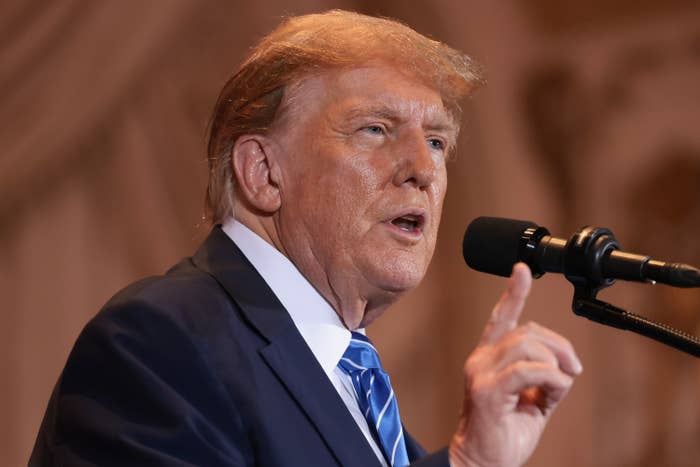 Trump in a suit and tie speaks into a microphone, gesturing with his hand, likely at a public or formal event