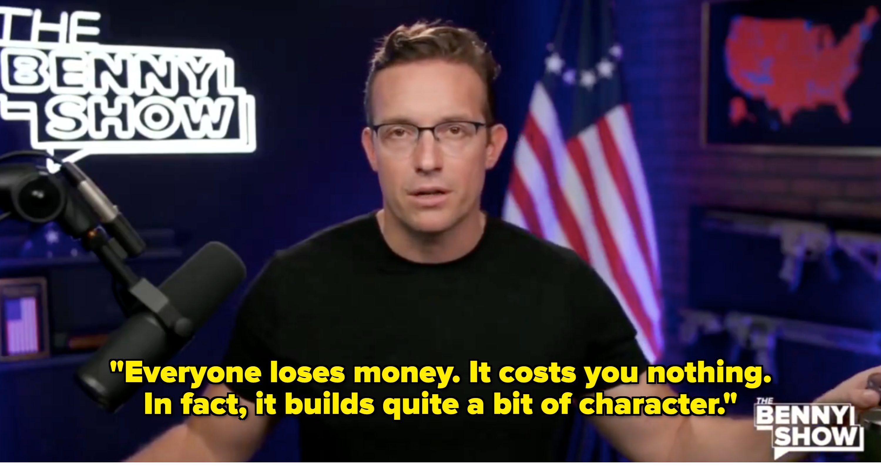 Benny Johnson wearing glasses and a black shirt speaks into a microphone on "The Benny Show" set with a U.S. flag in the background