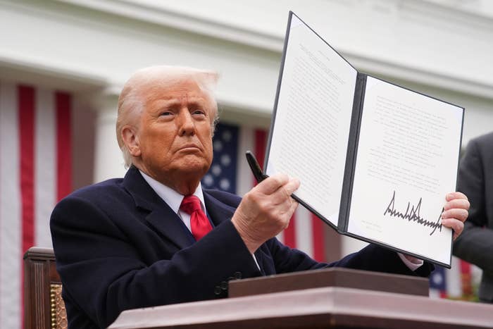Person in suit holding up a signed document with visible text, standing in front of American flags