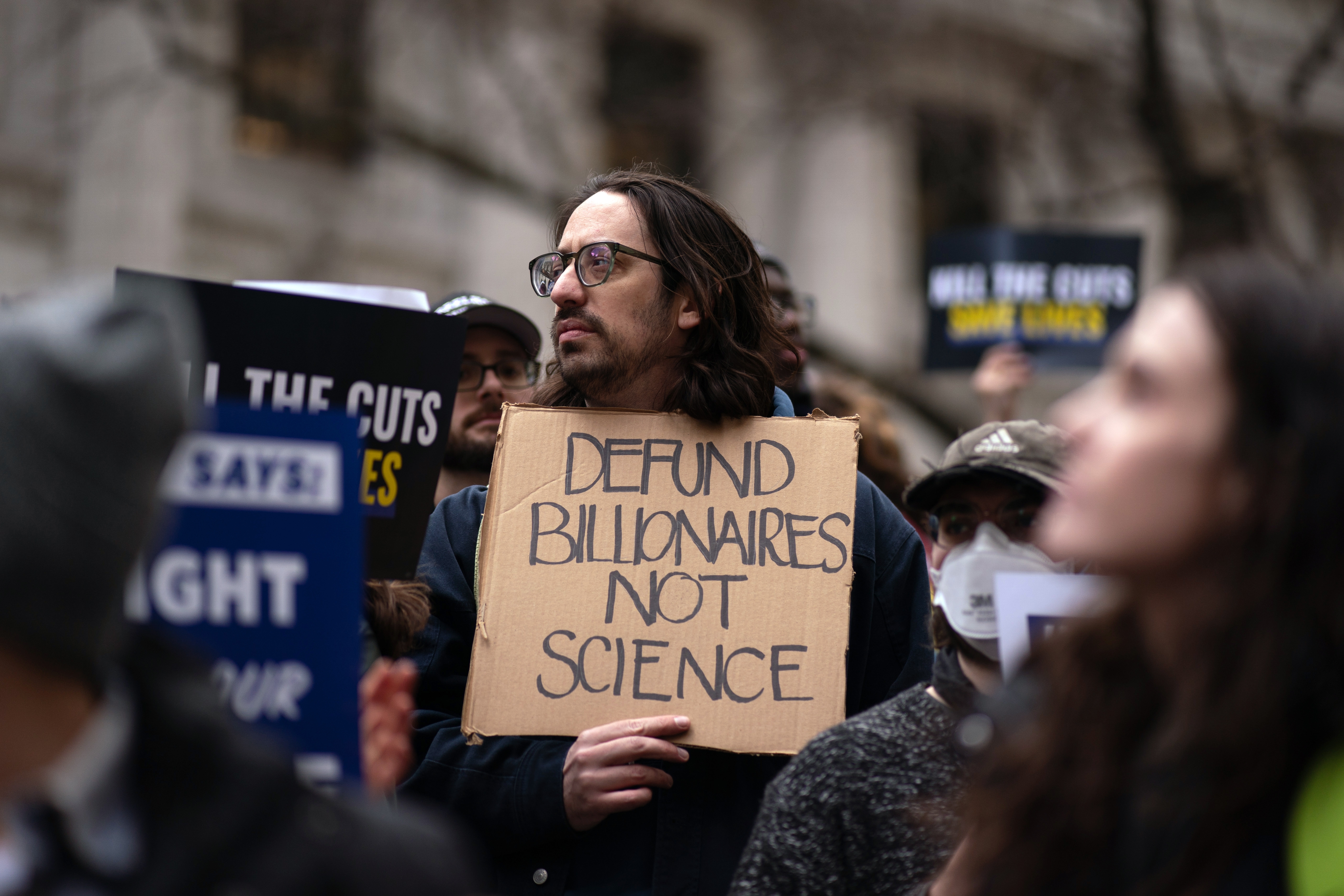 A man in a crowd holds a sign reading "Defund Billionaires Not Science" during a protest. Other signs are visible but not fully readable