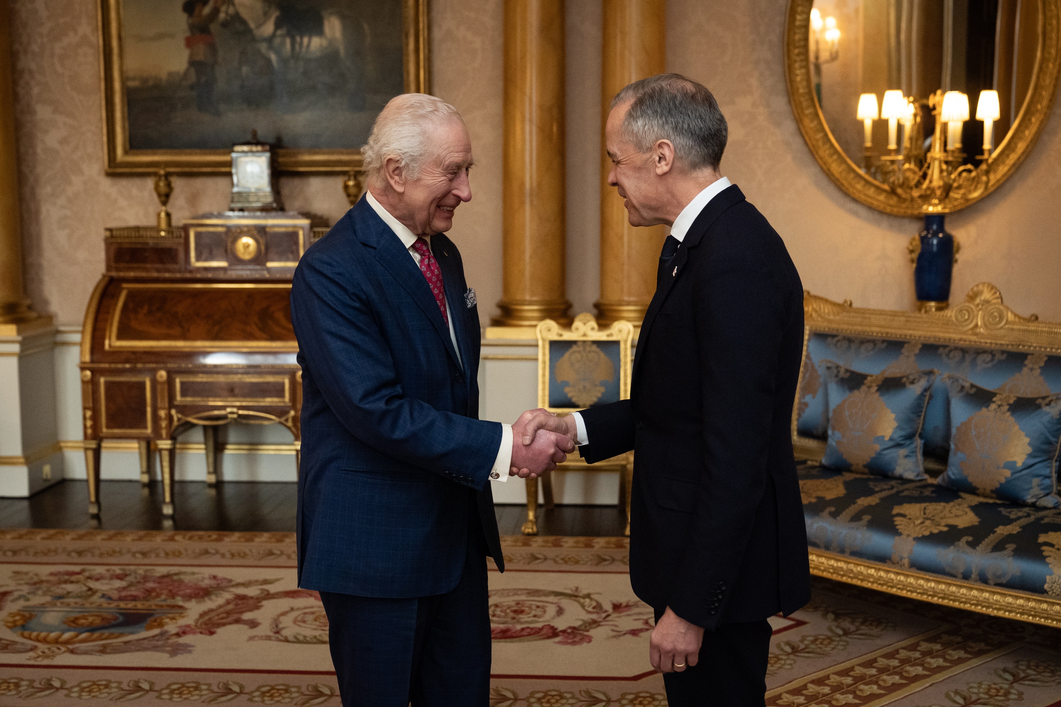 Two men in formal suits shake hands in an elegant room with ornate furniture and decor