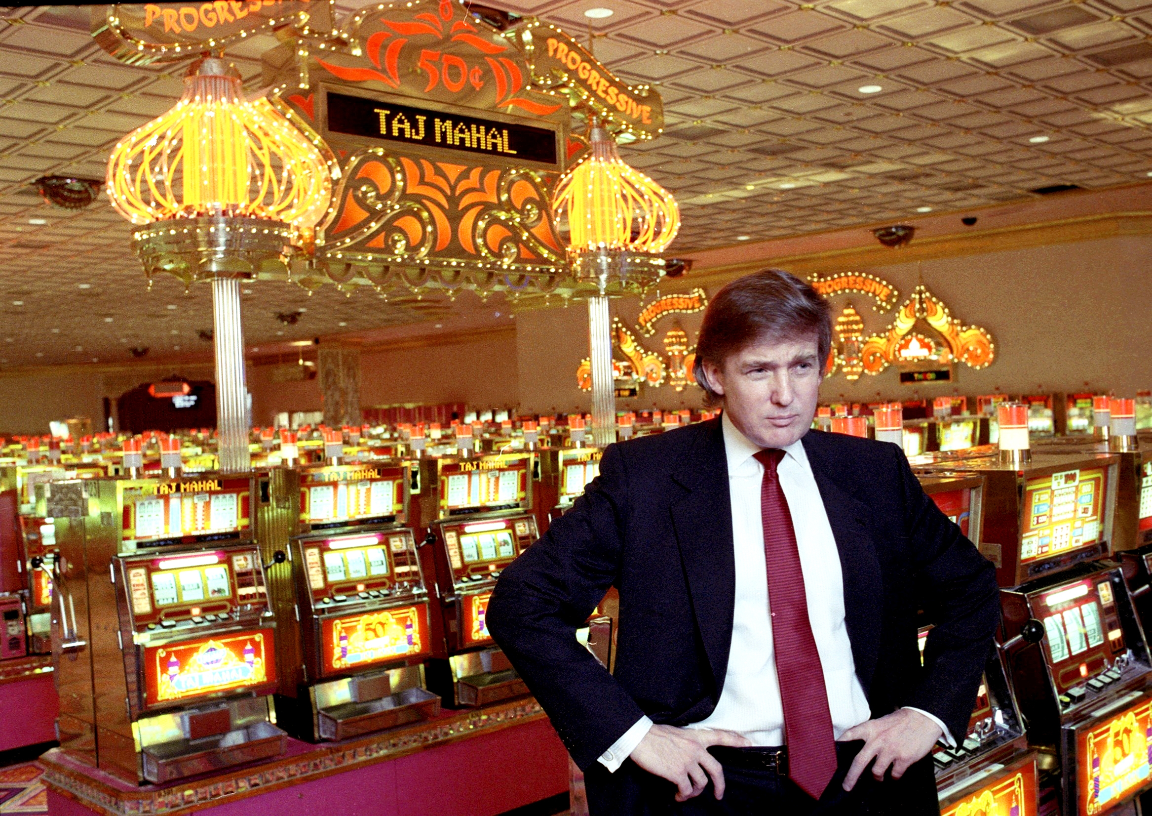 A person in a suit stands confidently in a casino filled with slot machines. The decor is ornate, featuring a prominent "Taj Mahal" sign