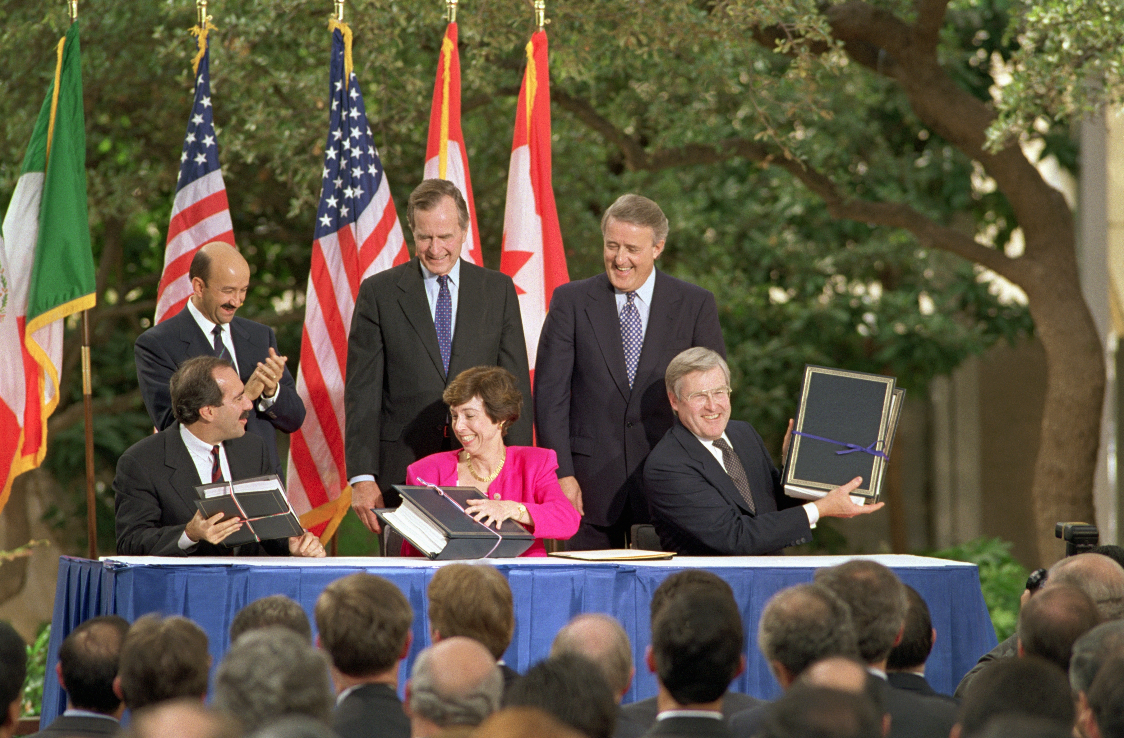 Five officials at a signing ceremony outdoors, each holding documents. Flags of various nations in the background