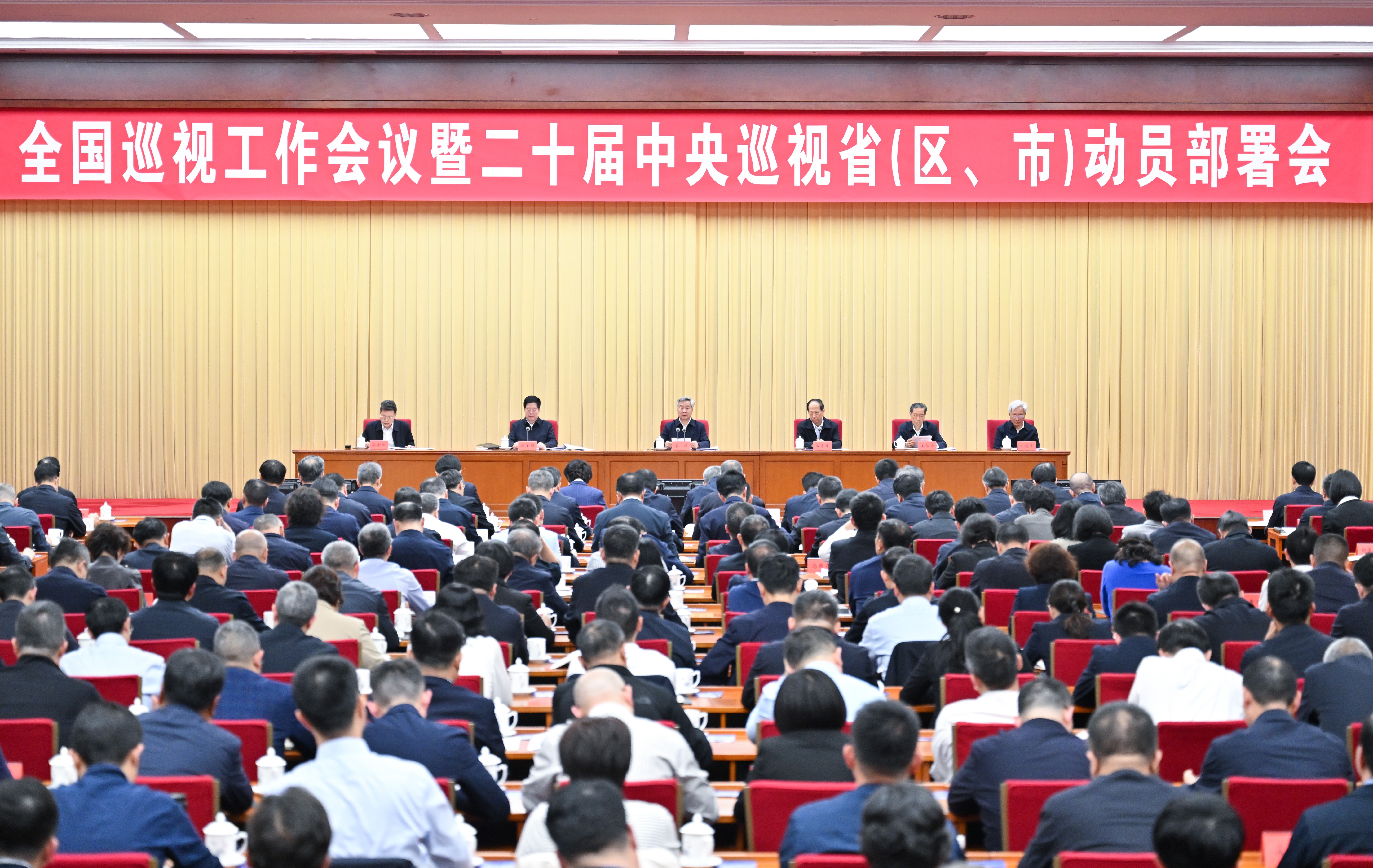 Large conference with officials seated at a long table on stage, addressing a crowd of suited attendees