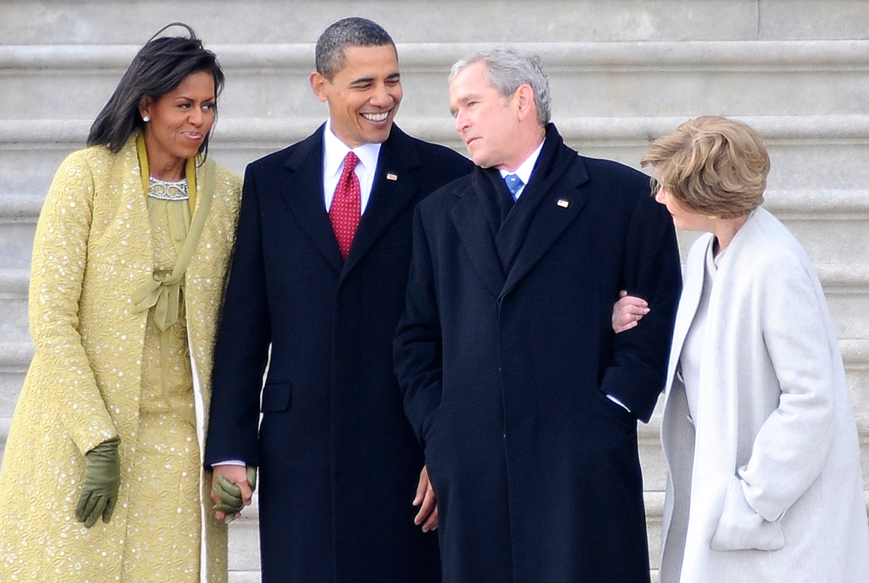 Four individuals stand together, smiling and conversing, dressed in formal winter coats on a stone staircase