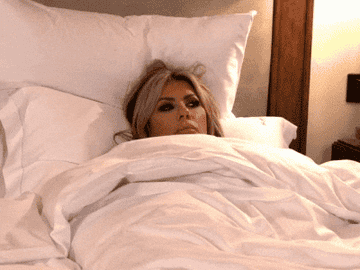 Person lying in bed, looking contemplative, head resting on a pillow, with a white comforter pulled up to their neck