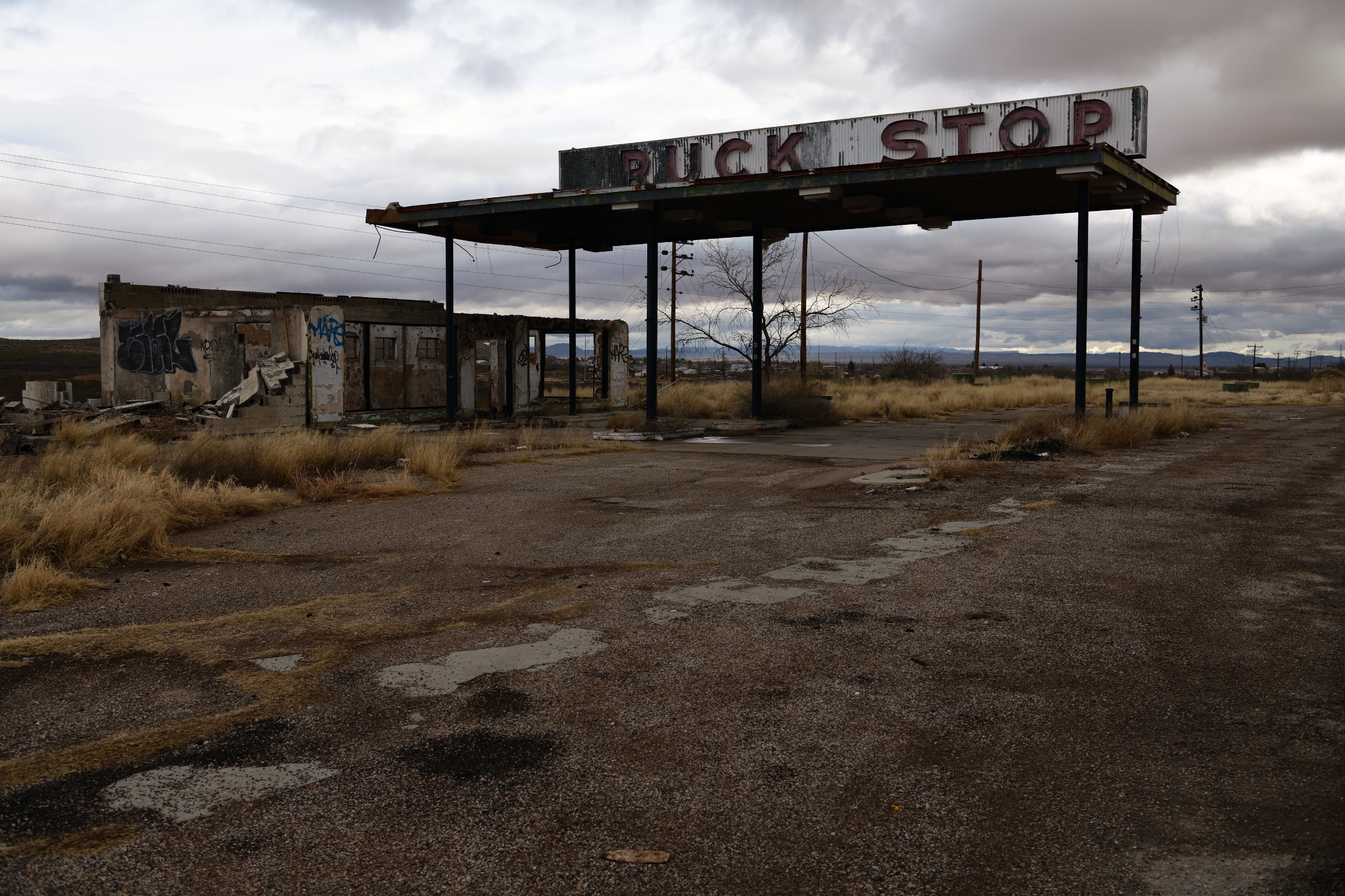 Abandoned gas station with a weathered canopy and deteriorating buildings in a deserted area. Signs of neglect and overgrown grass are visible