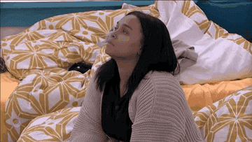 A person sitting on a bed with geometric-patterned bedding, looking up thoughtfully, wearing a cozy cardigan over a dark shirt