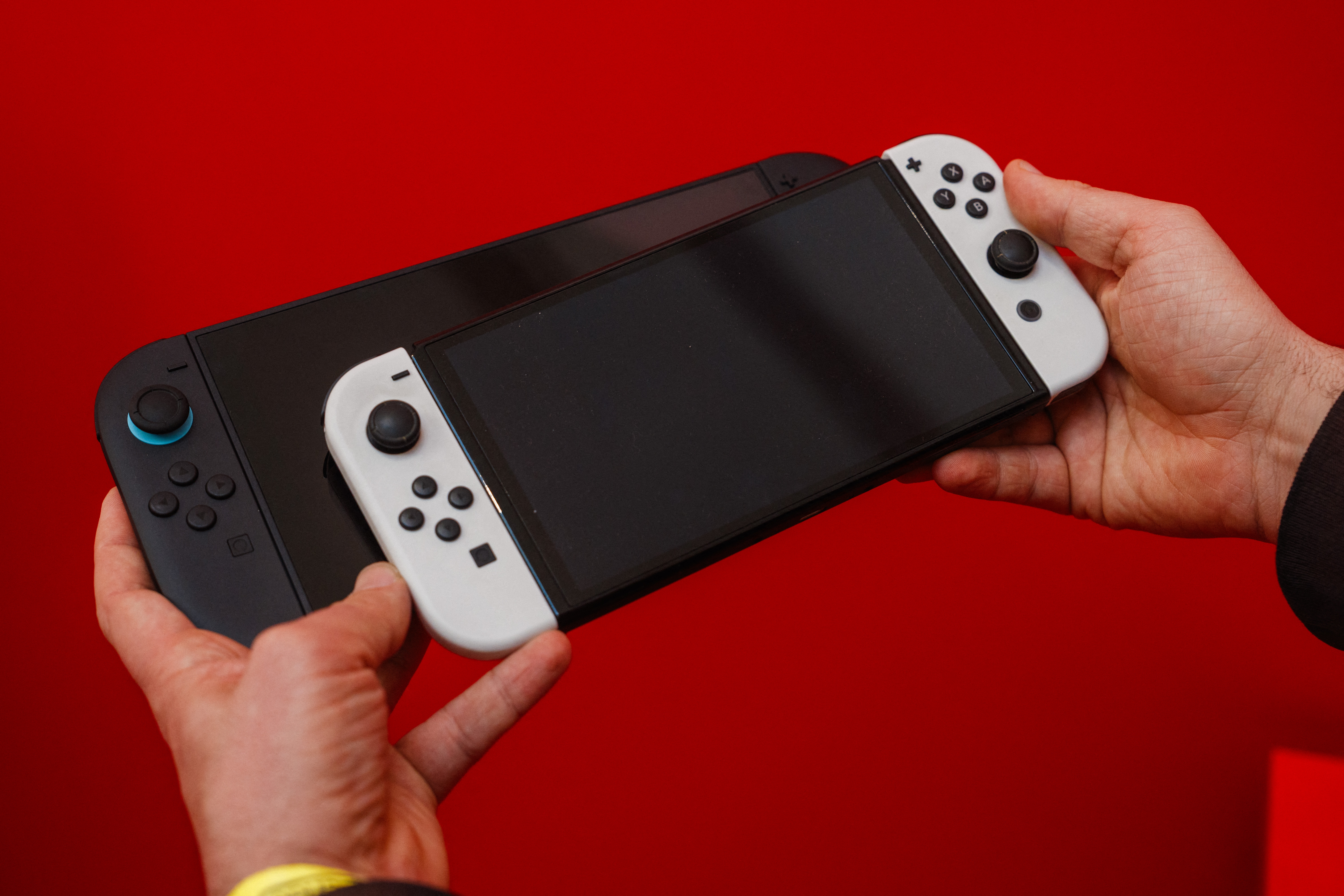 Two gaming consoles, with one person holding a larger black one and another person holding a smaller one with white controllers, set against a red backdrop