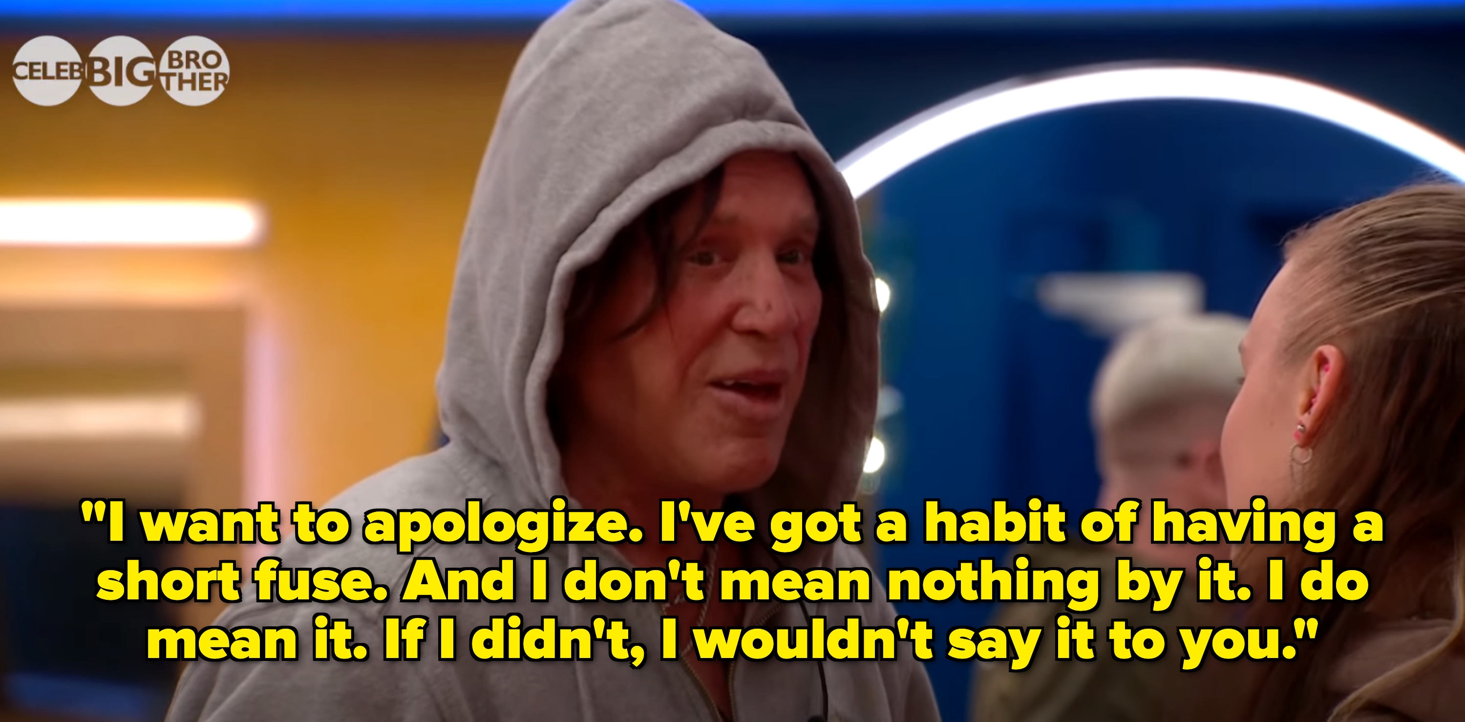 Person in a gray hoodie speaking to someone off-camera in a room with bright lights, likely a reality TV setting