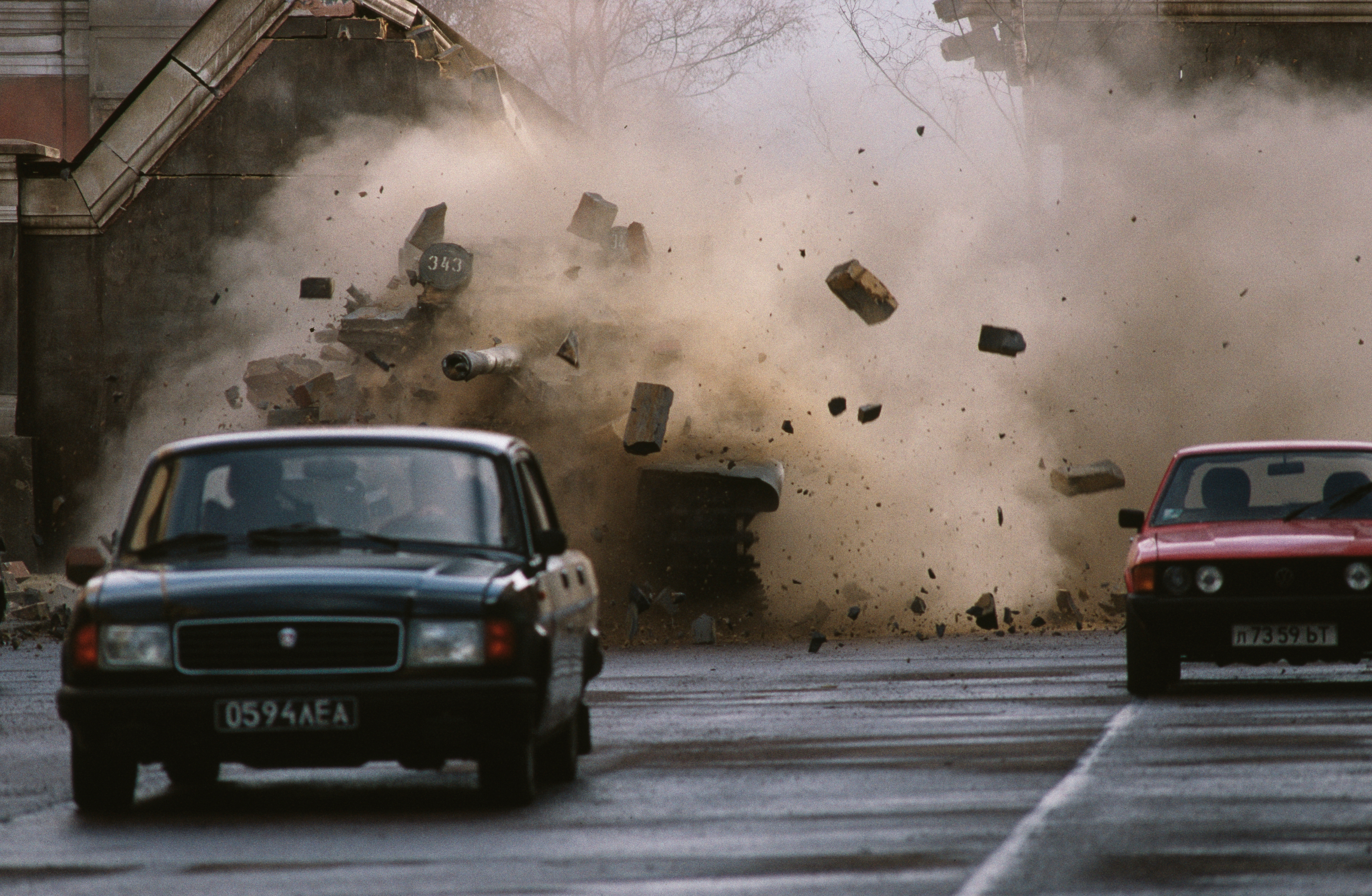 Debris and dust fill the street as a building wall collapses, while cars drive past the scene