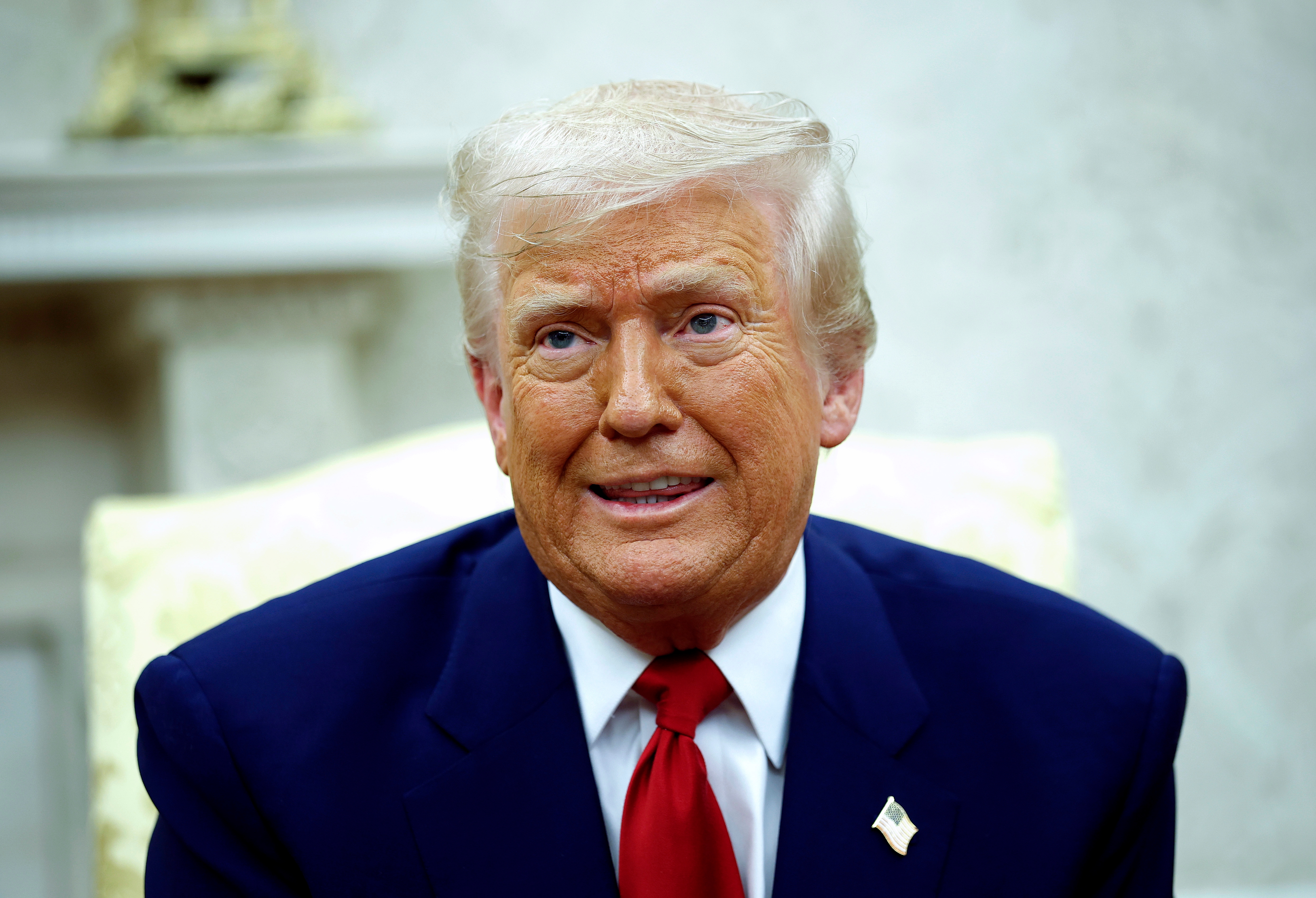 A person in a suit and red tie sits indoors, with a slight smile, in front of a blurred light-colored background