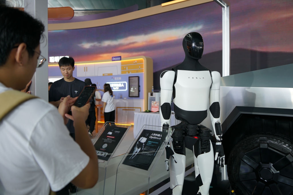 A person takes a photo of a display featuring a humanoid robot at a technology event; other attendees and exhibits are visible