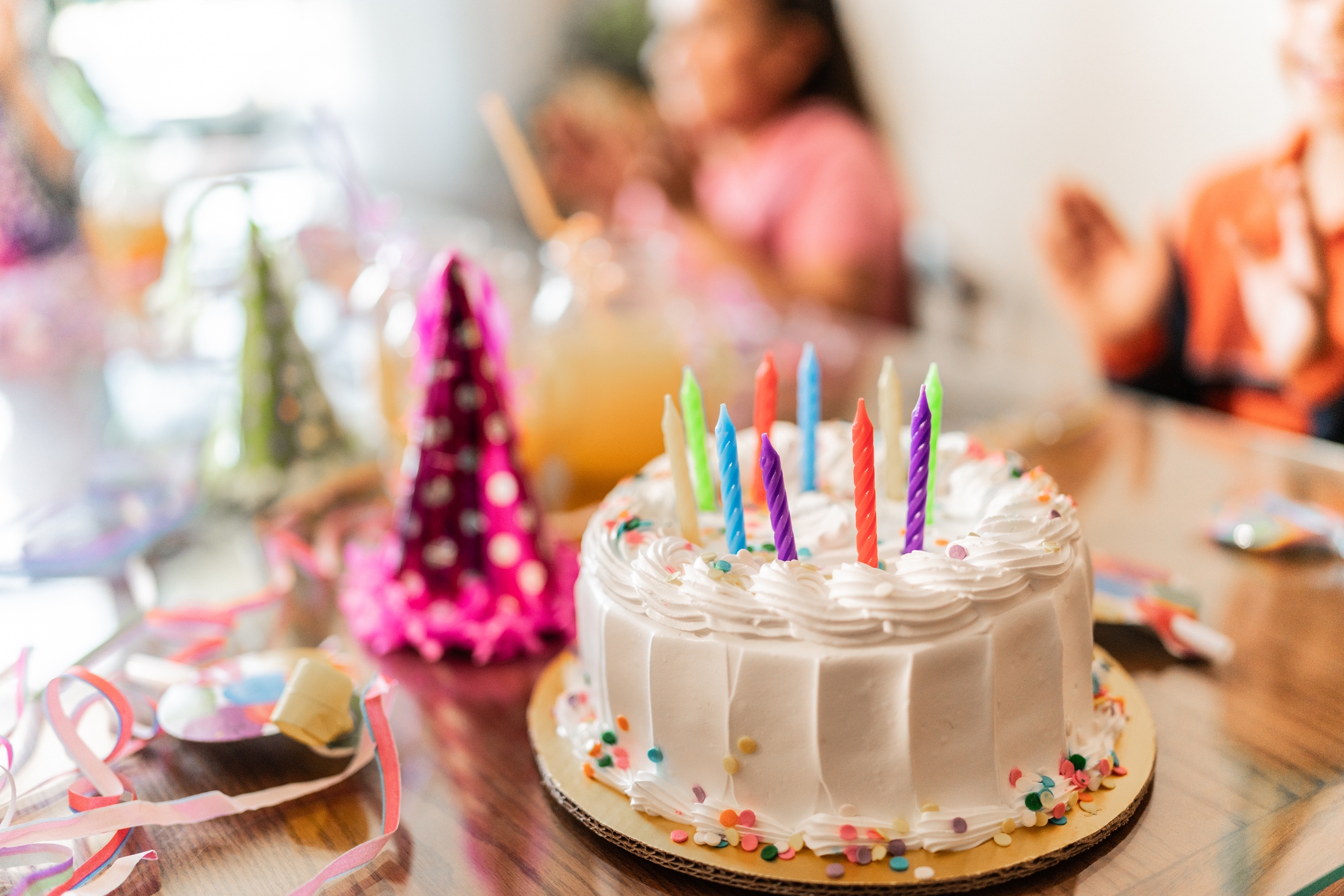 A festive birthday scene with a cake topped with colorful candles, surrounded by party hats and decorations. People are seen blurred in the background