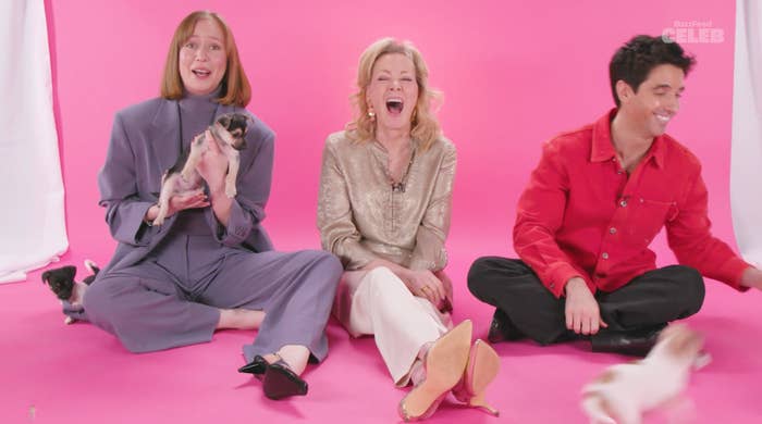 Three people seated on the floor, holding playful puppies, laughing joyfully. The person on the left wears a loose suit; the center wears a shiny top; the right wears a casual jacket