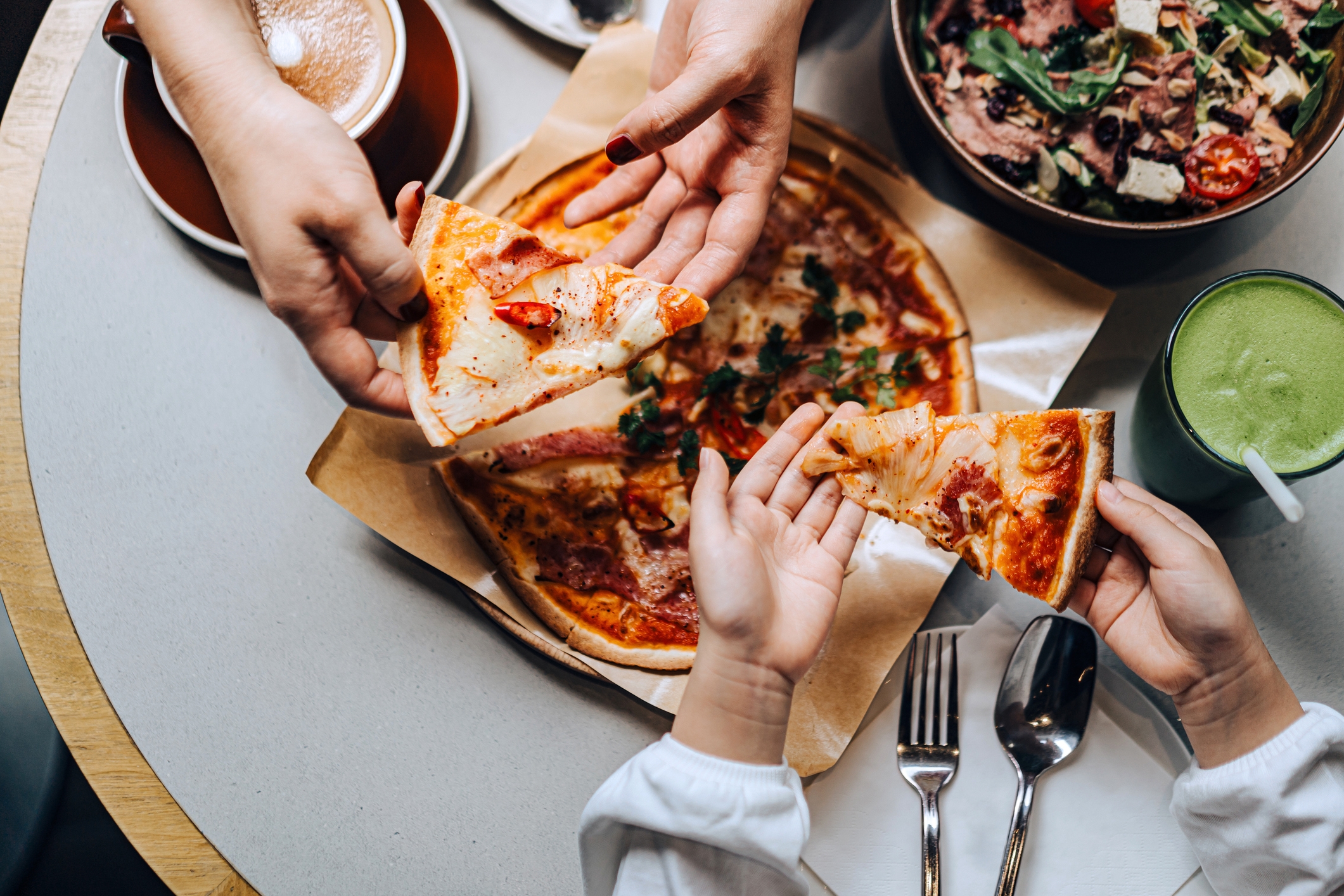 Three people share a pizza at a table, each holding a slice. A salad and green smoothie are also on the table