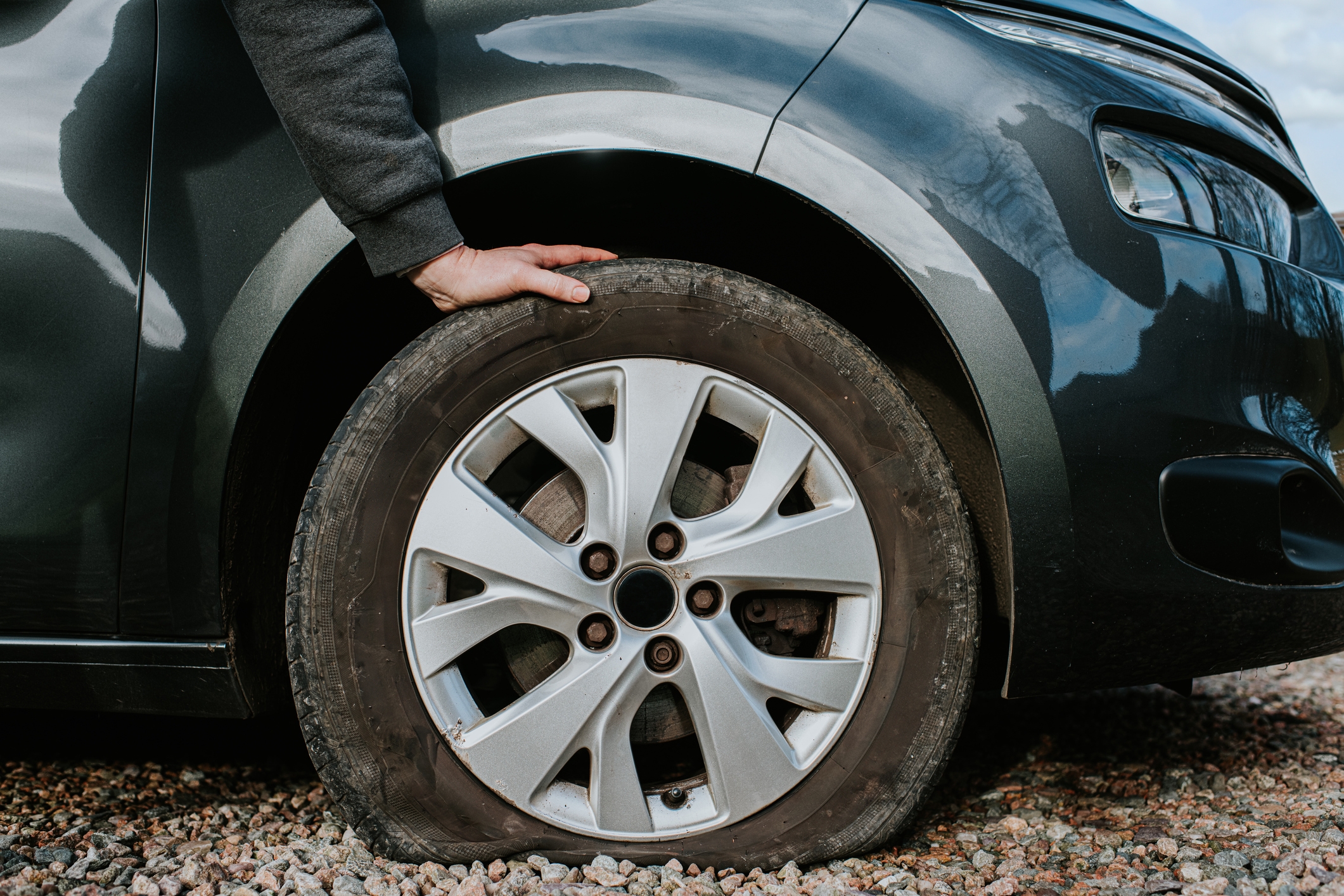 A person examines a flat tire on a parked car over gravel