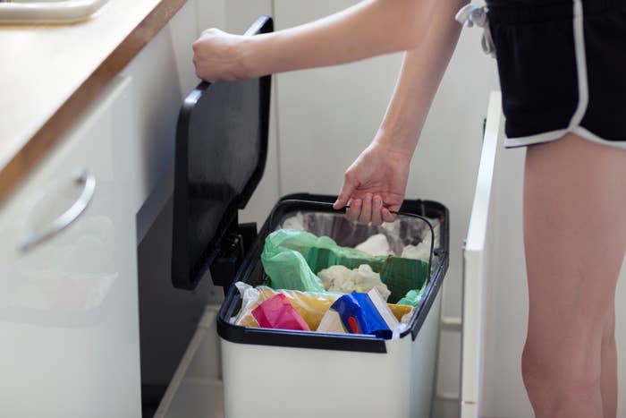 A person opens a kitchen trash bin filled with various colored bags and waste, showing a household setting