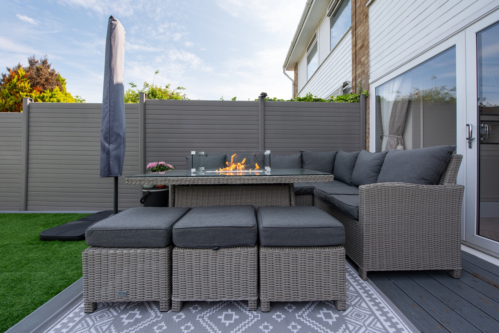 Outdoor patio with wicker furniture, a corner couch, ottomans, and a table with a rectangular fire pit. Fence and sliding doors in the background