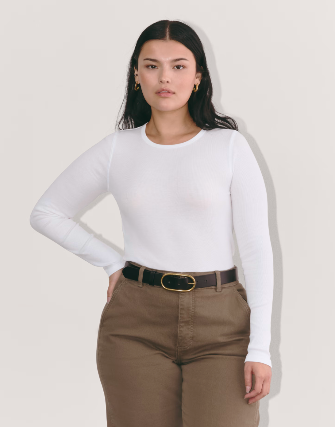 Person wearing a fitted white long-sleeve top and brown pants with a black belt. Gold hoop earrings visible