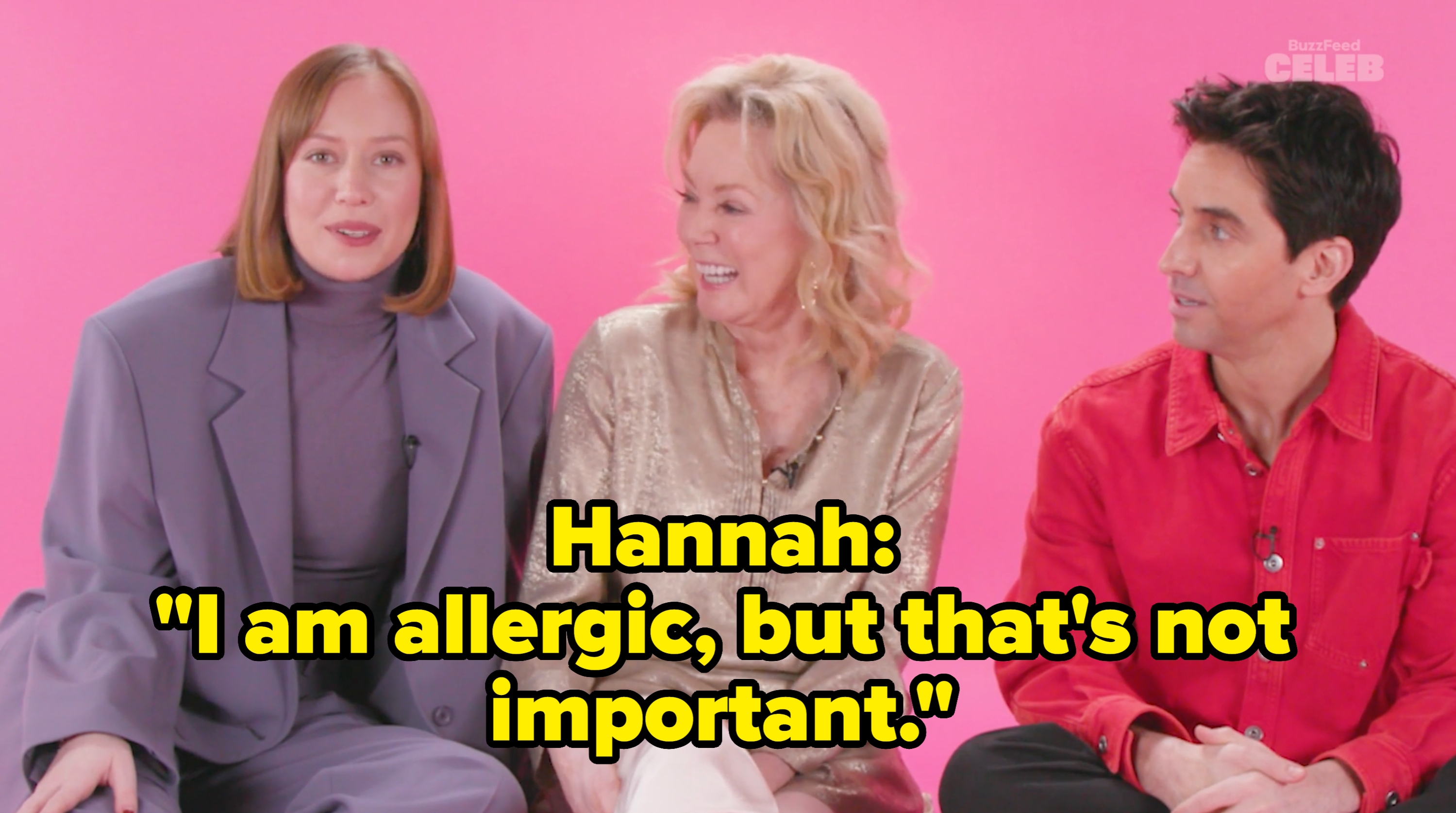 Three people sit together smiling on a set. One wears a suit, another is in a shimmery blouse, and the third person wears a casual shirt