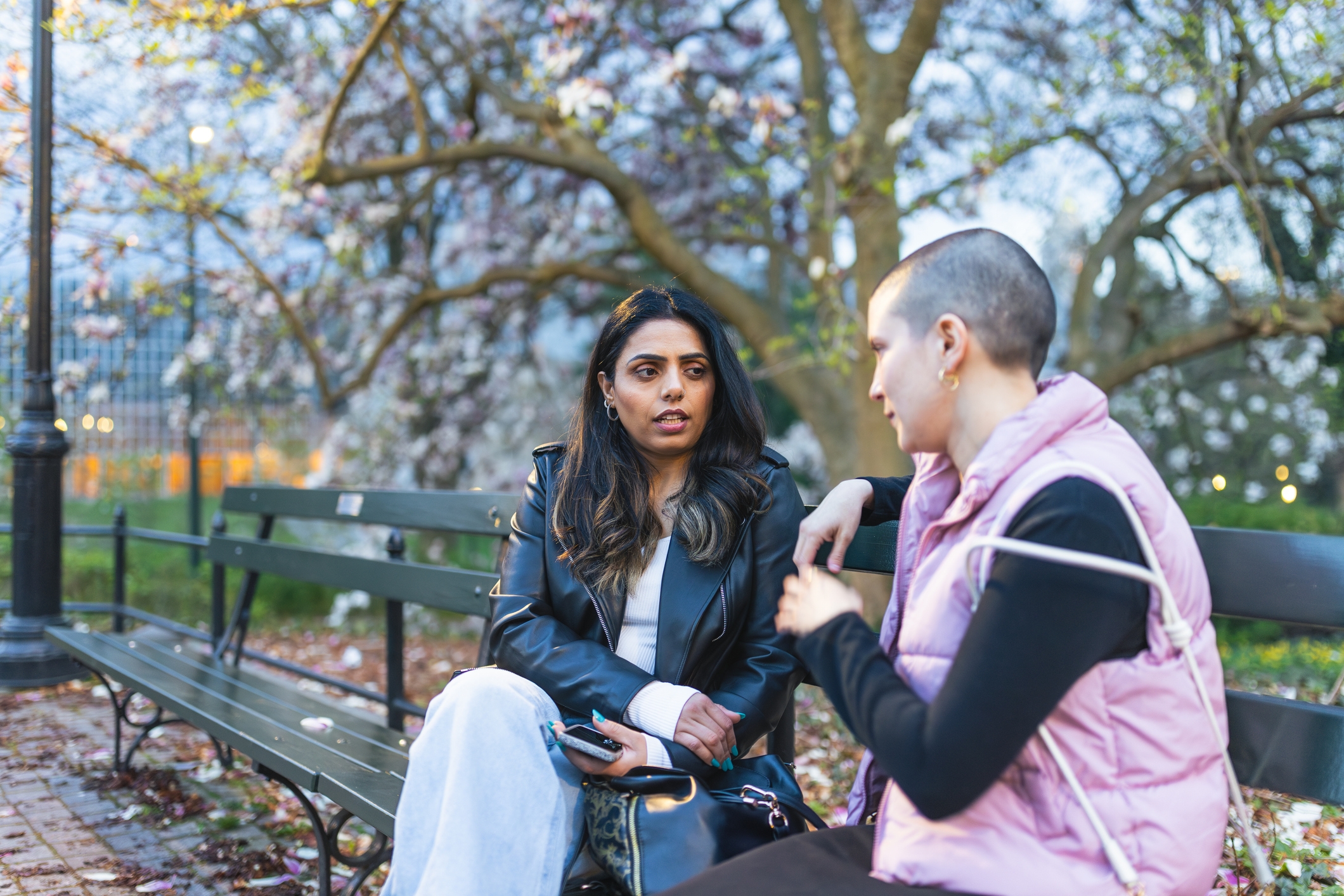 Two people sitting on a park bench having a conversation; one is wearing a leather jacket, and the other a quilted vest