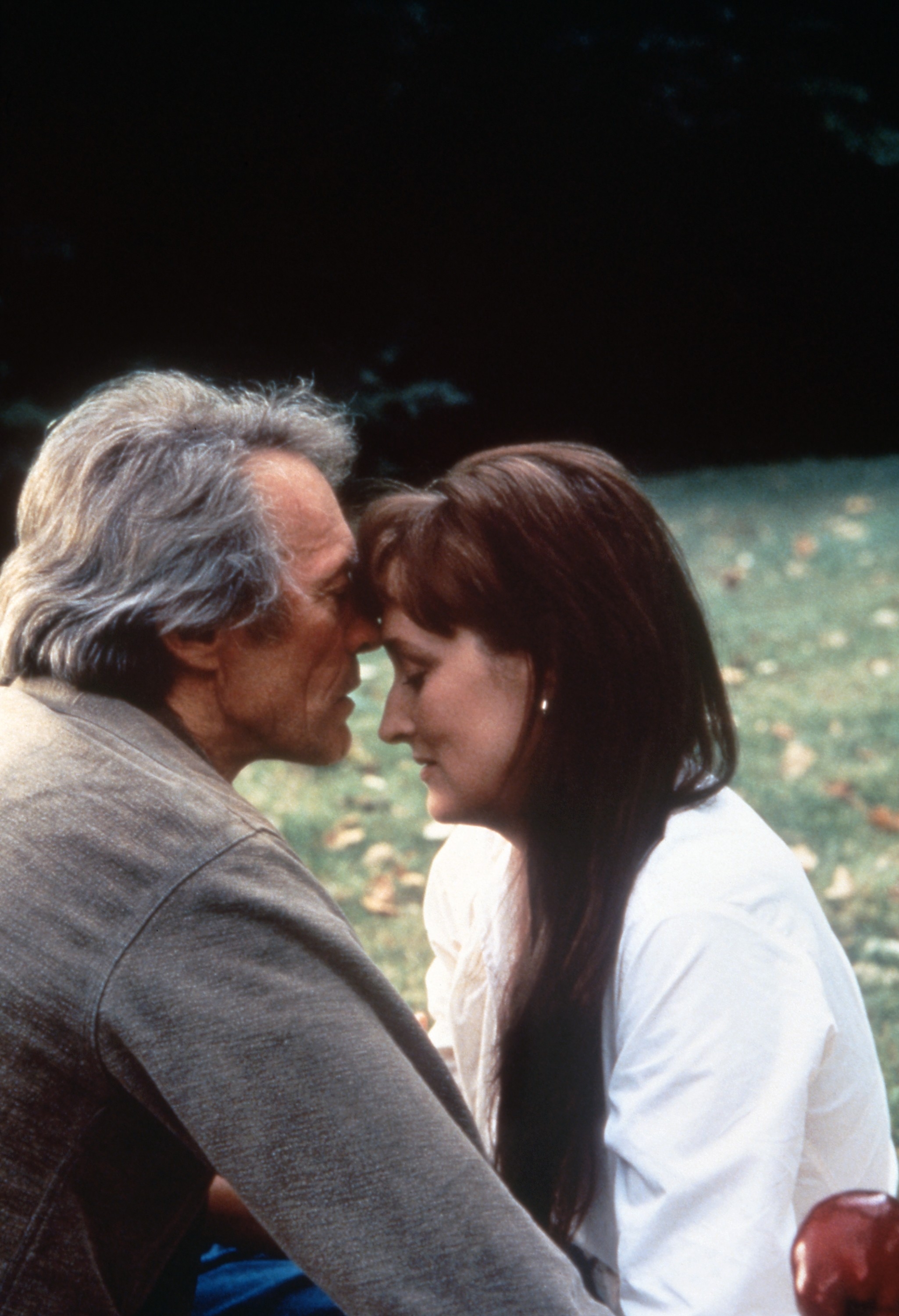 An elderly man and woman sit closely in an outdoor setting, touching foreheads in a tender, intimate moment