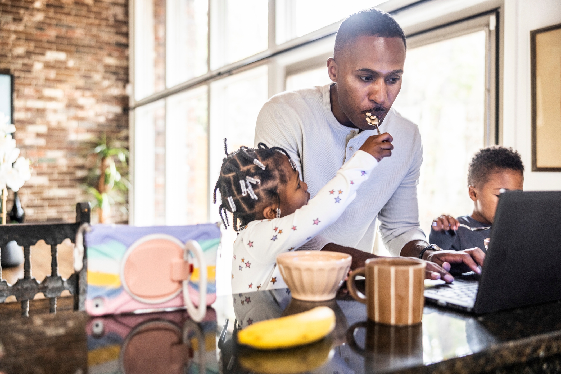 Parent multitasks on a laptop while two children eat breakfast at a kitchen counter
