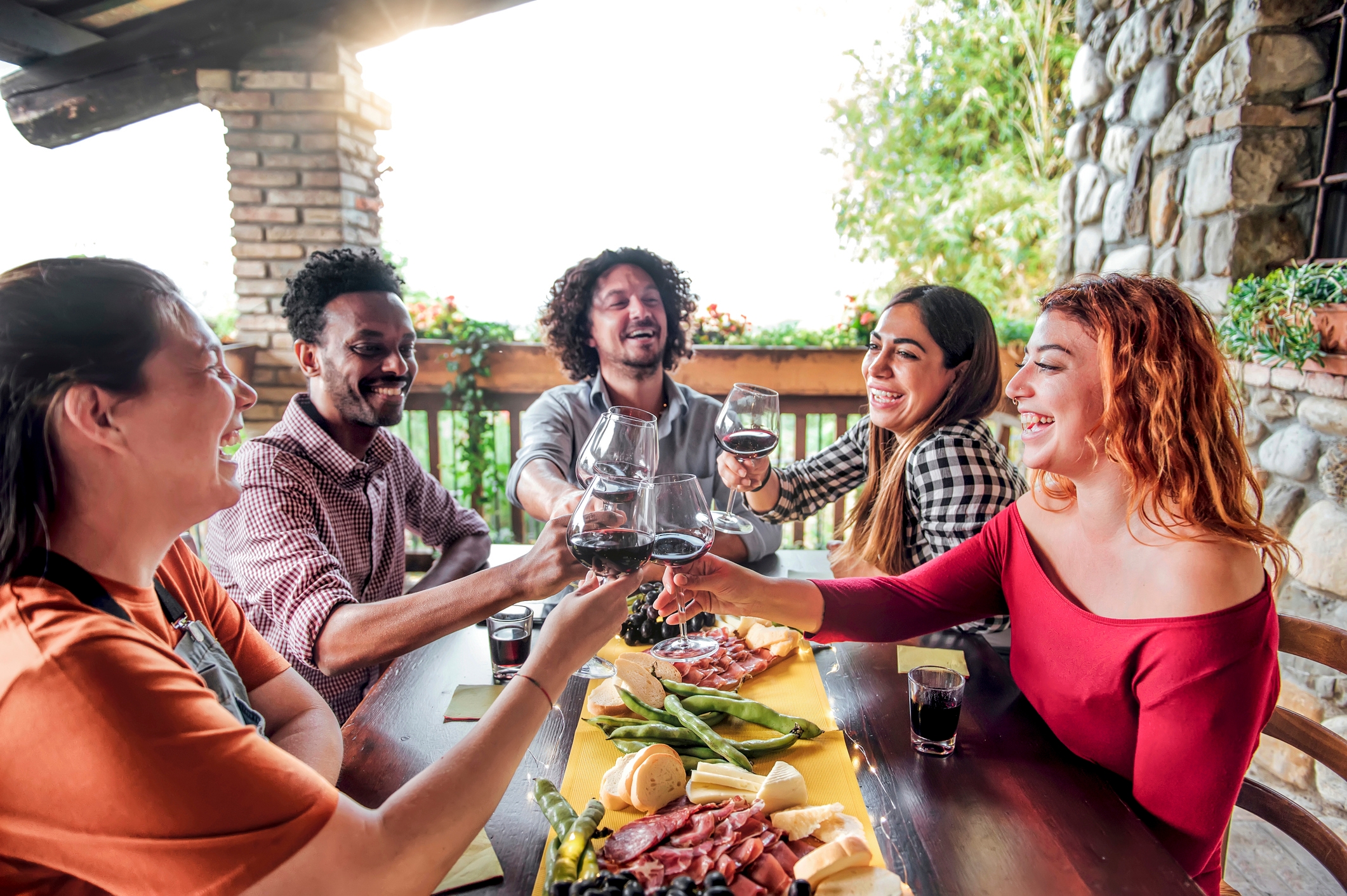 Group of people sitting at an outdoor table, laughing and toasting with wine glasses over a meal. Rustic stone and wood setting