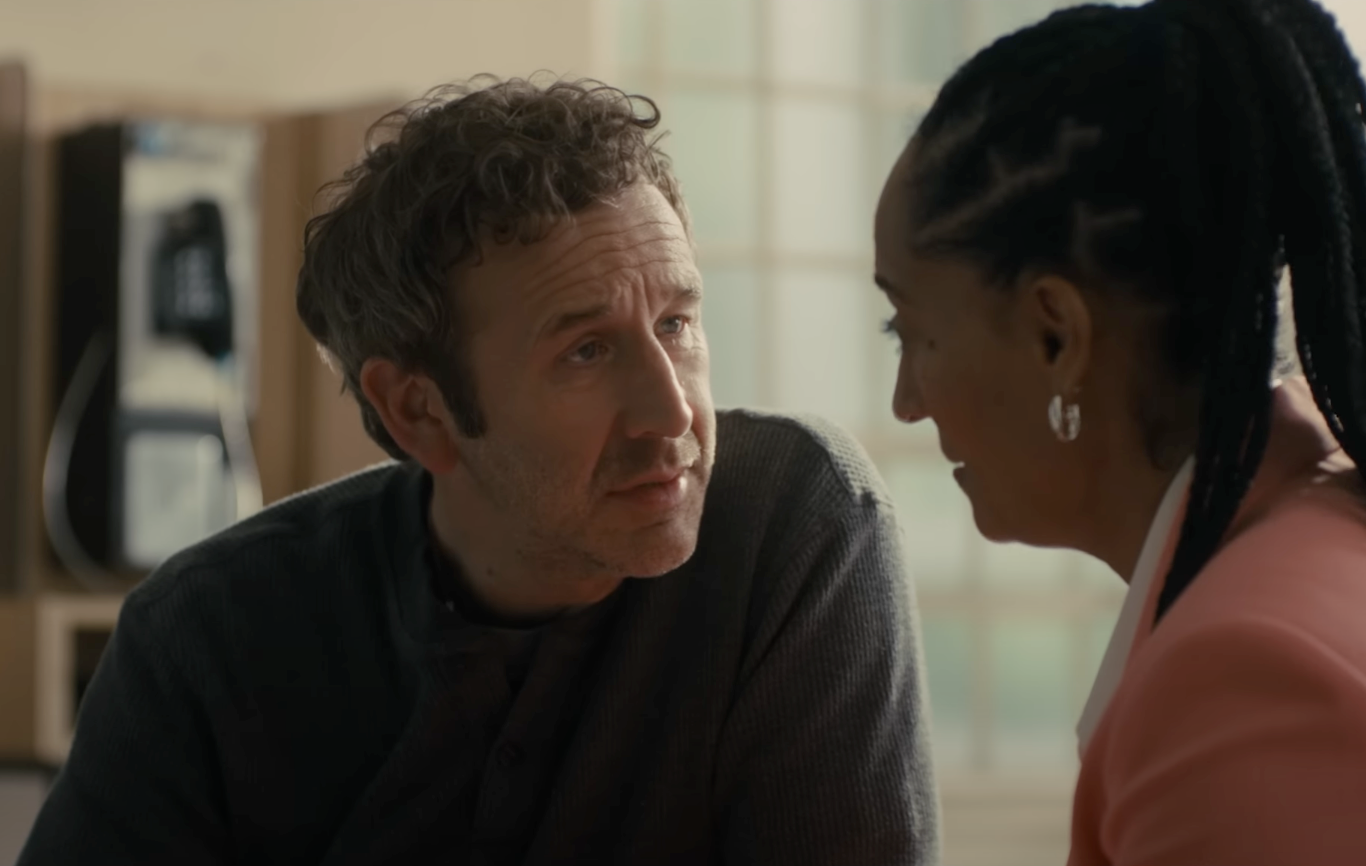 Two people sit closely, engaged in a conversation. The man wears a sweater, the woman has braided hair and wears a blazer. The setting appears casual