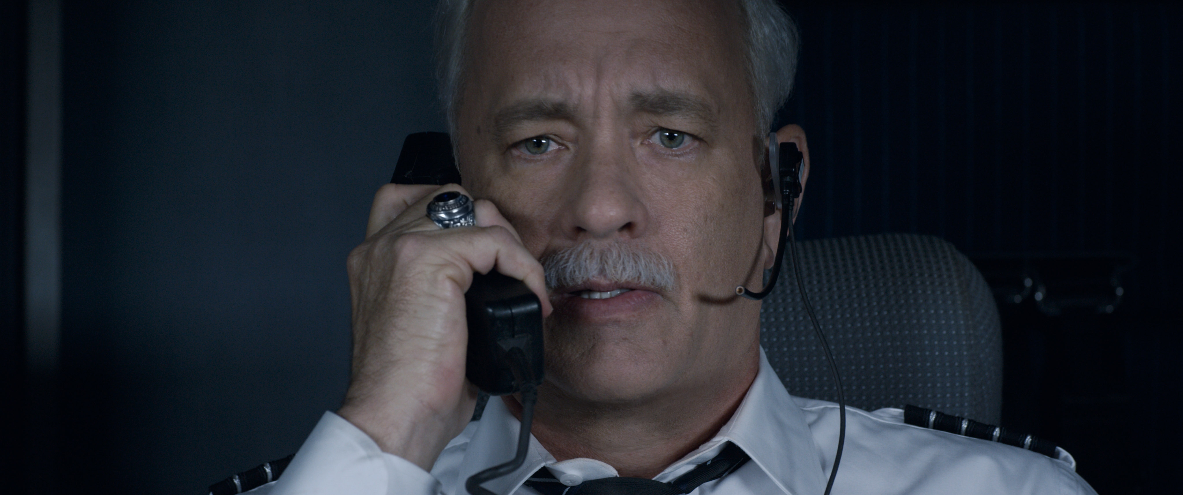 A person in a pilot uniform, wearing a headset, looks concerned while speaking on a phone