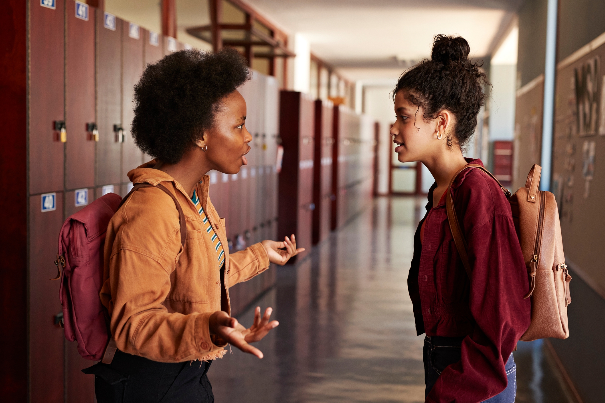 Two students are talking animatedly in a school hallway, both wearing backpacks and casual outfits. Lockers line the corridor