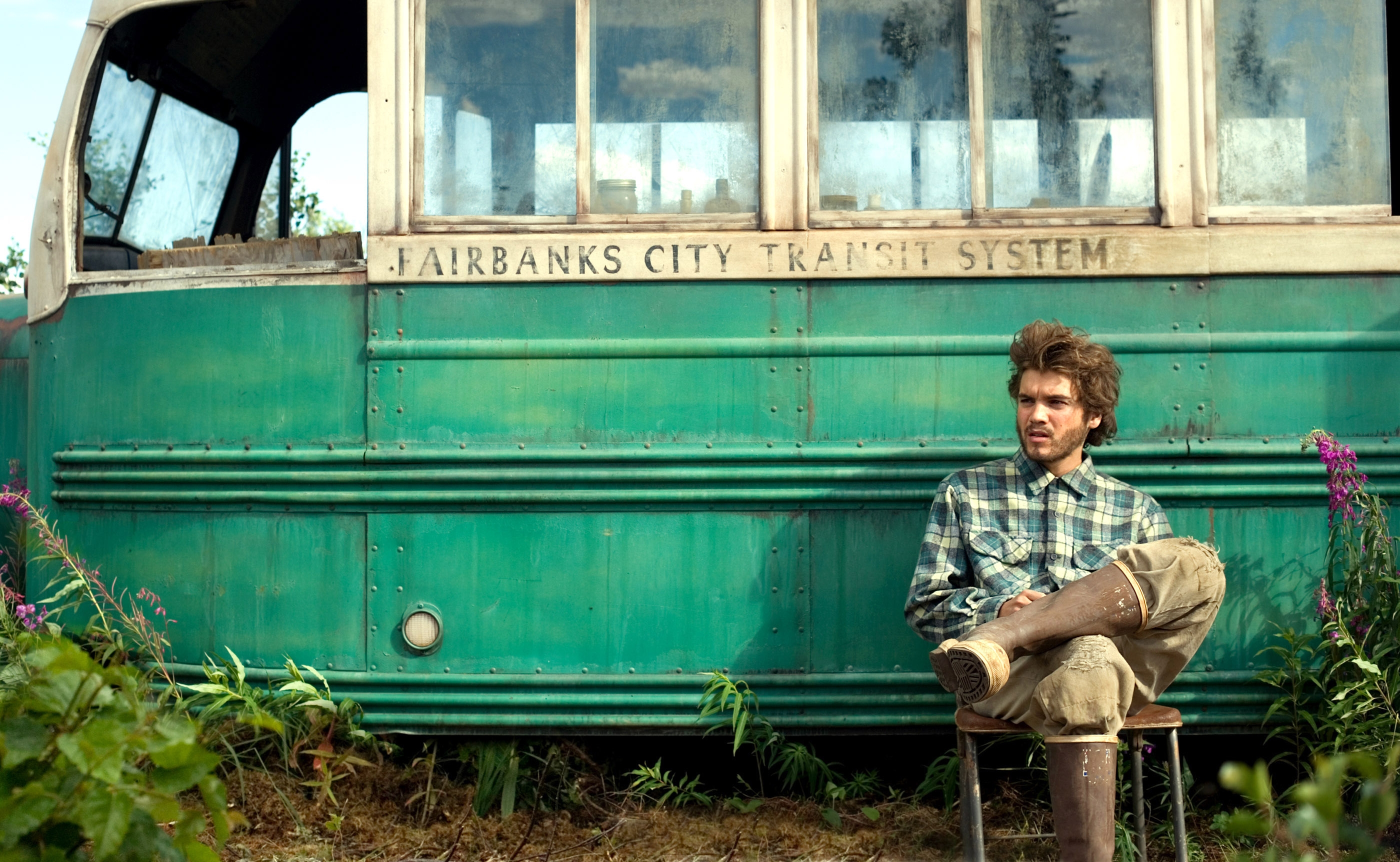 Person sitting on a stool beside an old, abandoned bus with &quot;Fairbanks City Transit System&quot; written on it