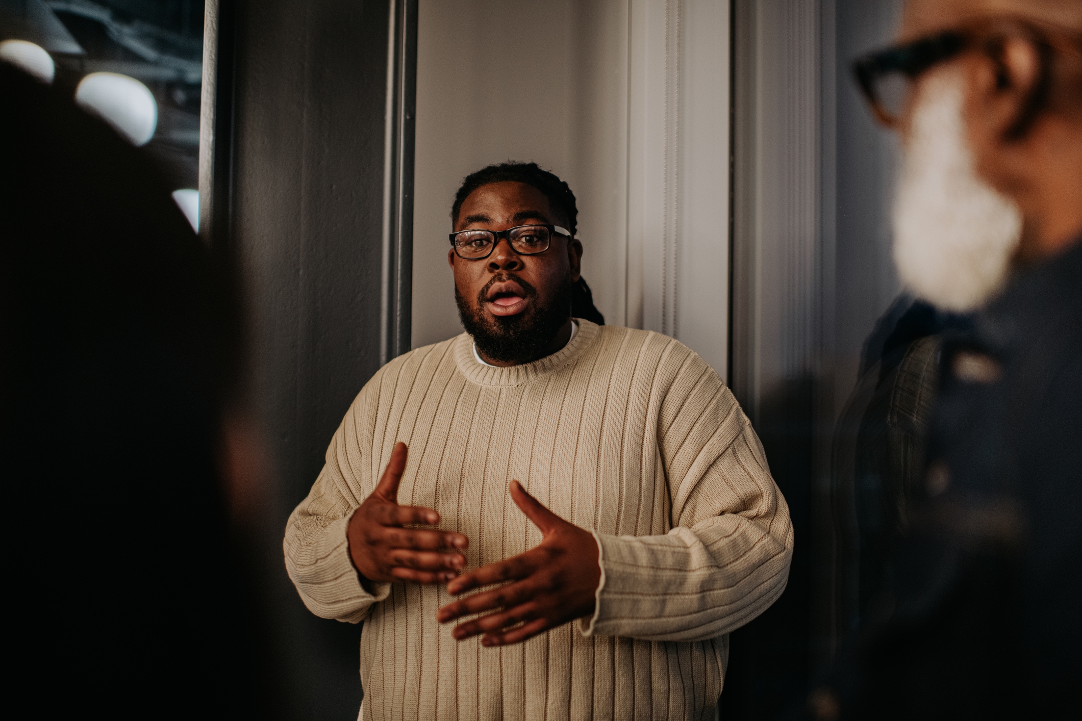 Person speaking passionately while gesturing with hands, wearing a ribbed sweater. Indoor setting with others partially visible in the foreground