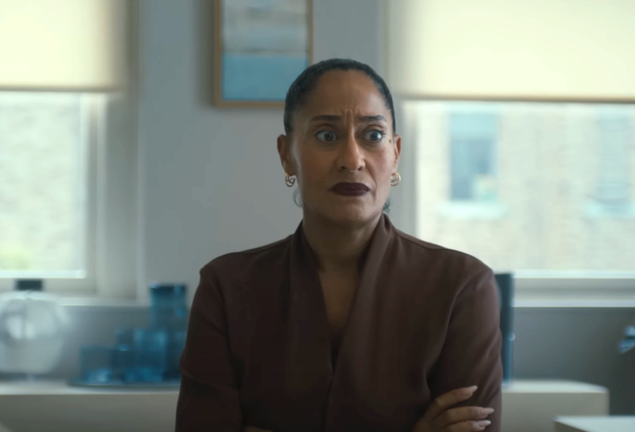 Person with an expressive look stands indoors with arms crossed, wearing a dark jacket