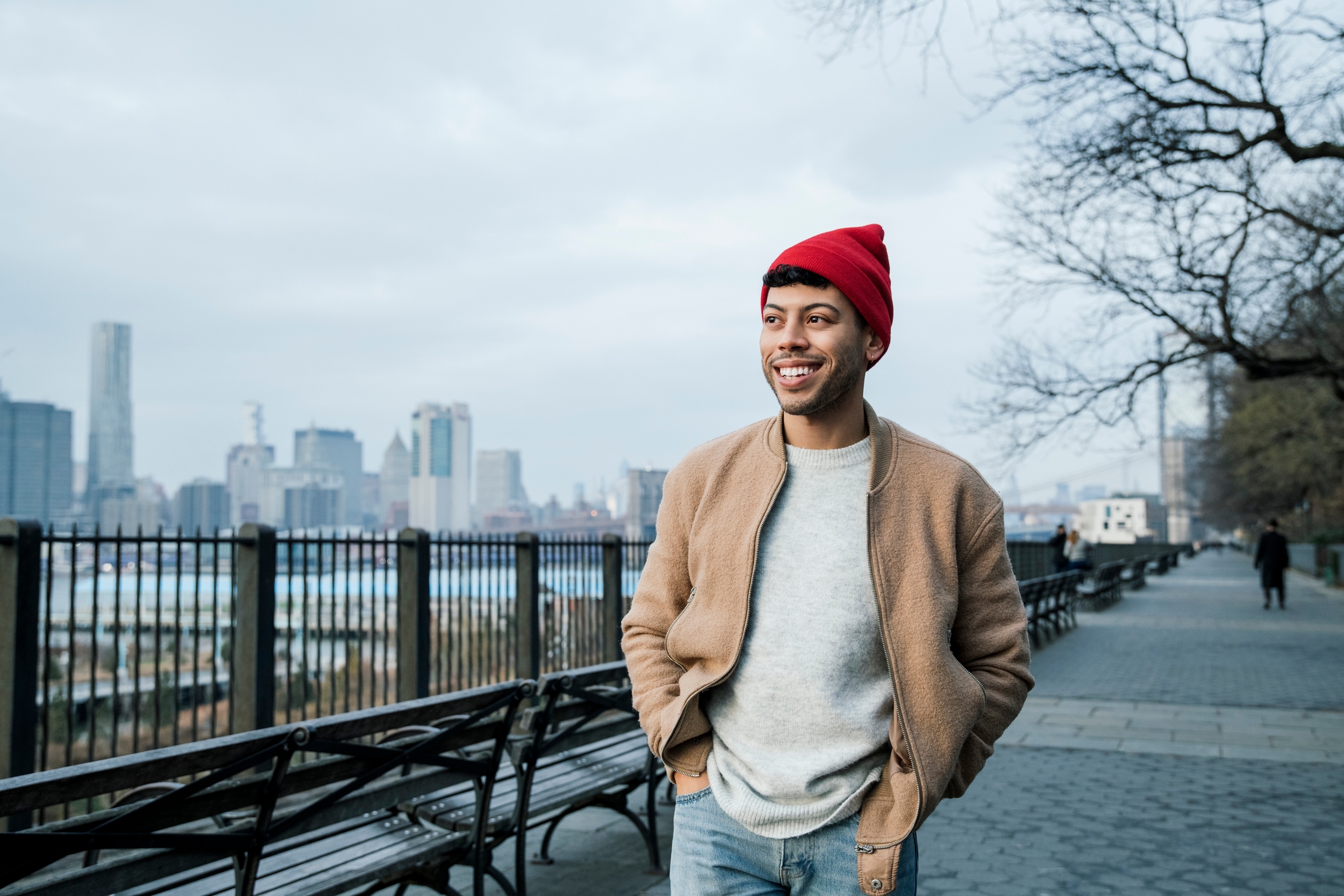 Person in casual attire with a red beanie smiles while walking on a city street with a skyline view in the background