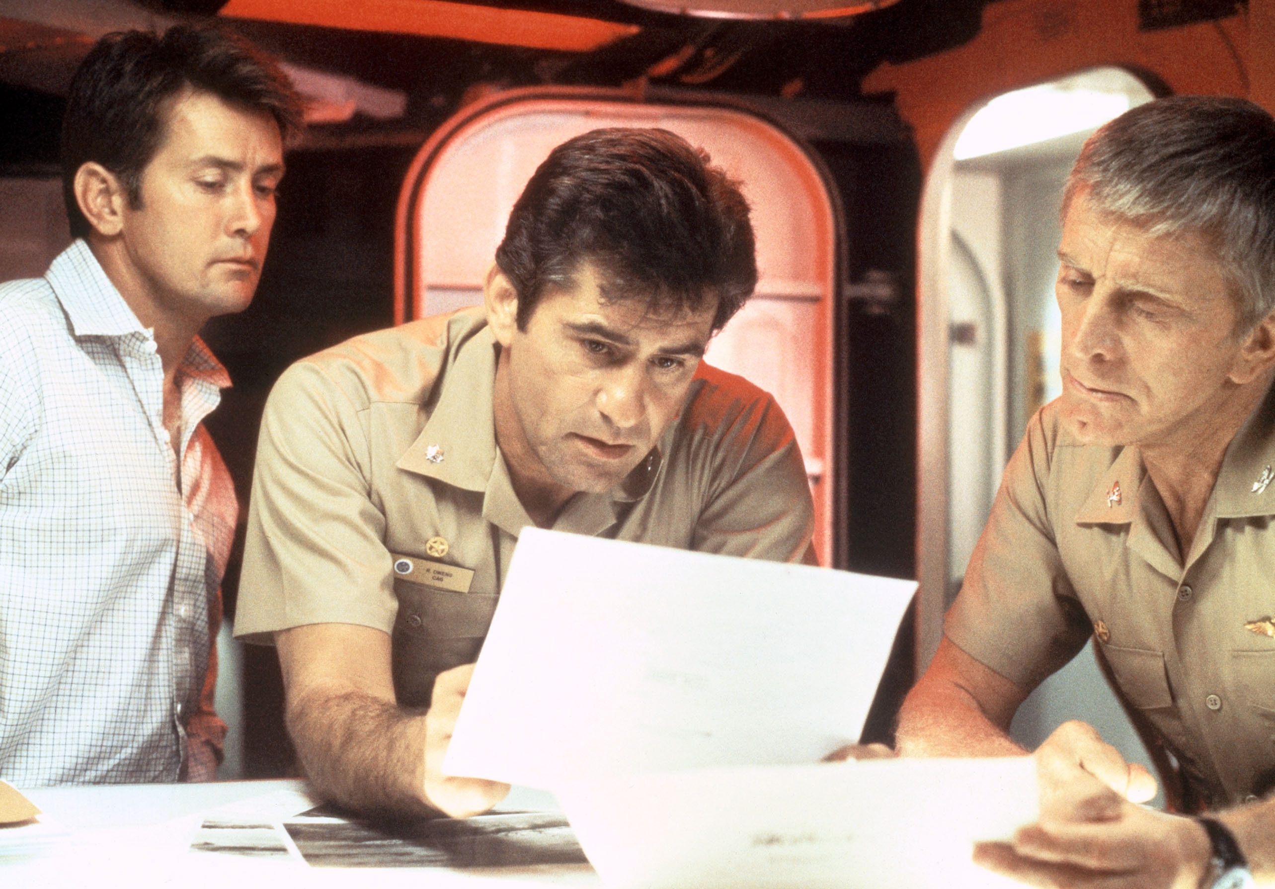 Three men in naval uniforms review documents intently at a table. They appear to be in a submarine setting