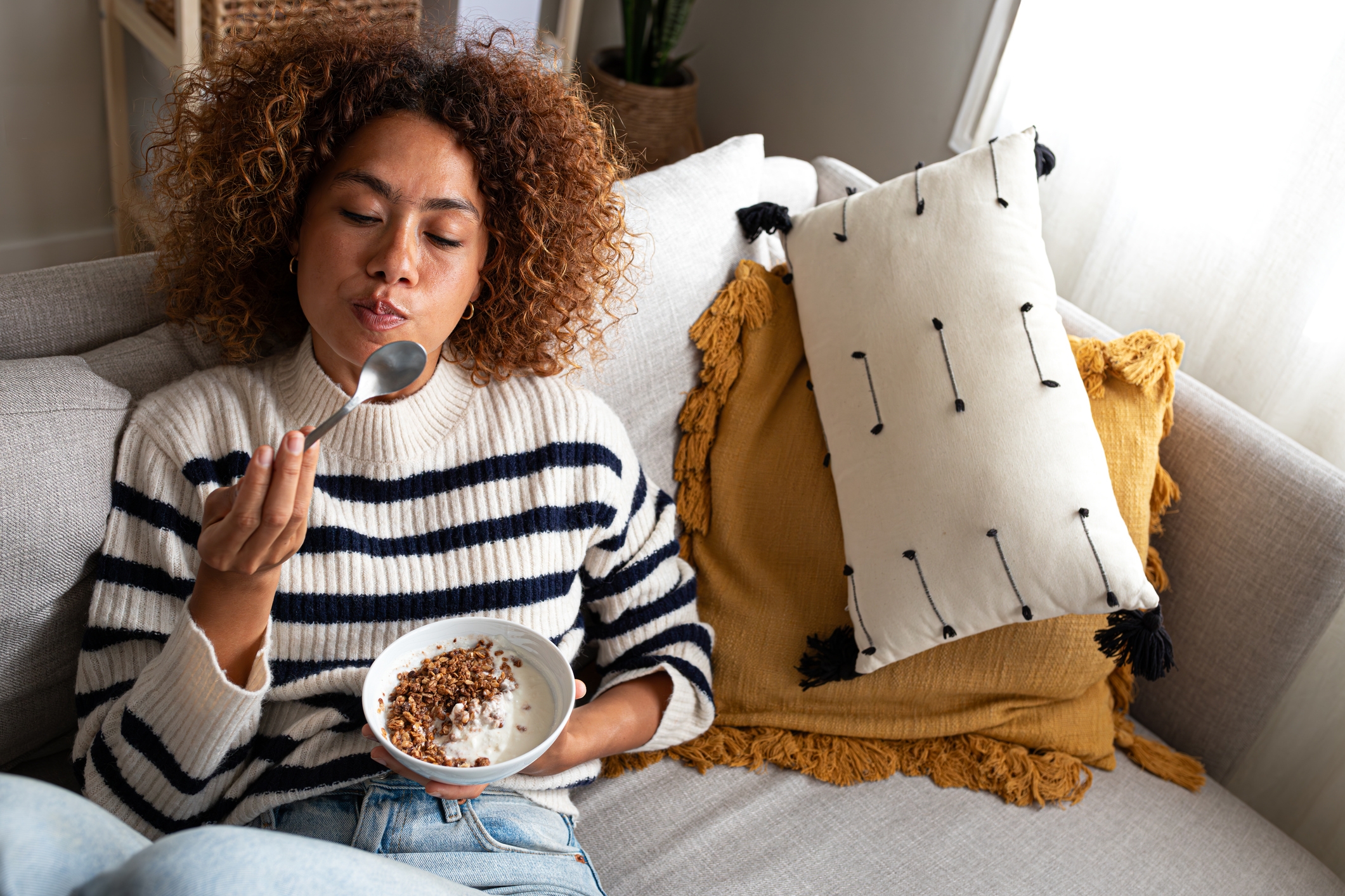 Person relaxing on a couch, wearing a striped sweater, eating from a bowl. Cozy pillows are in the background