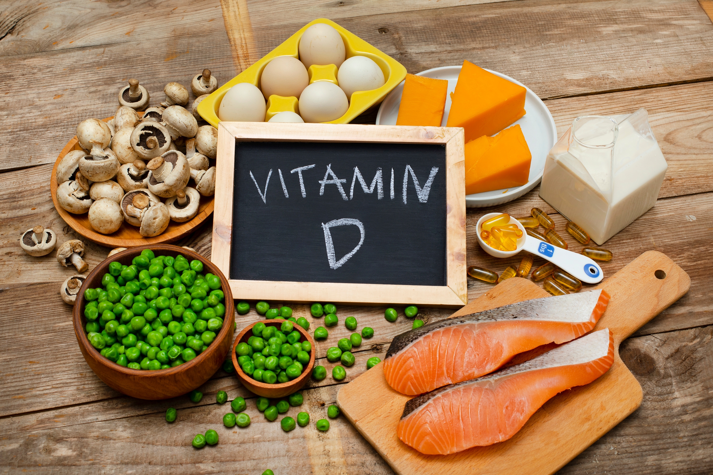 Fish, cheese, eggs, peas, and mushrooms arranged around a chalkboard labeled &quot;Vitamin D&quot; on a wooden table