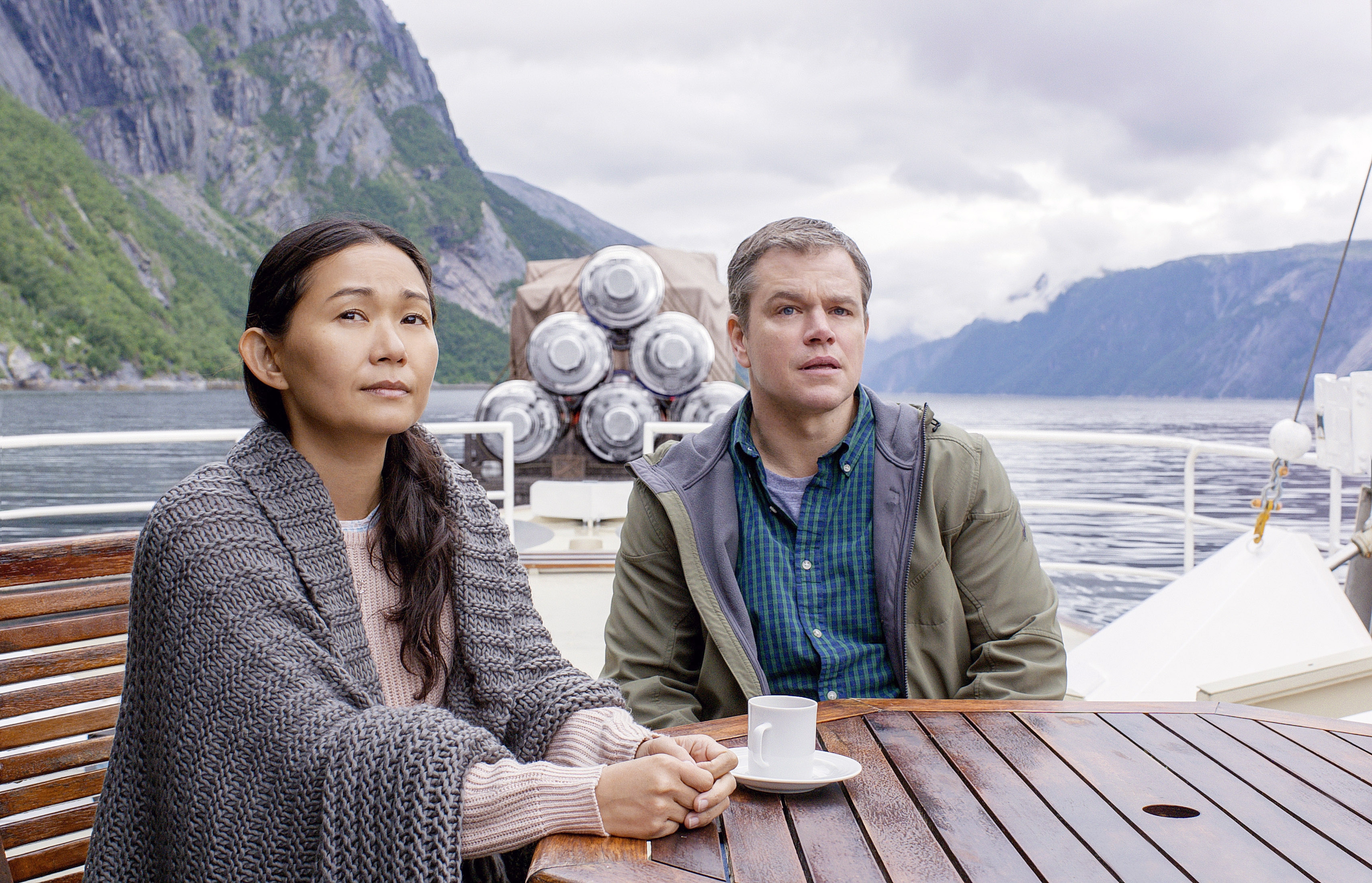Two people sit on a boat's deck with scenic mountain views in the background, one with a sweater and another with a jacket, both looking reflective