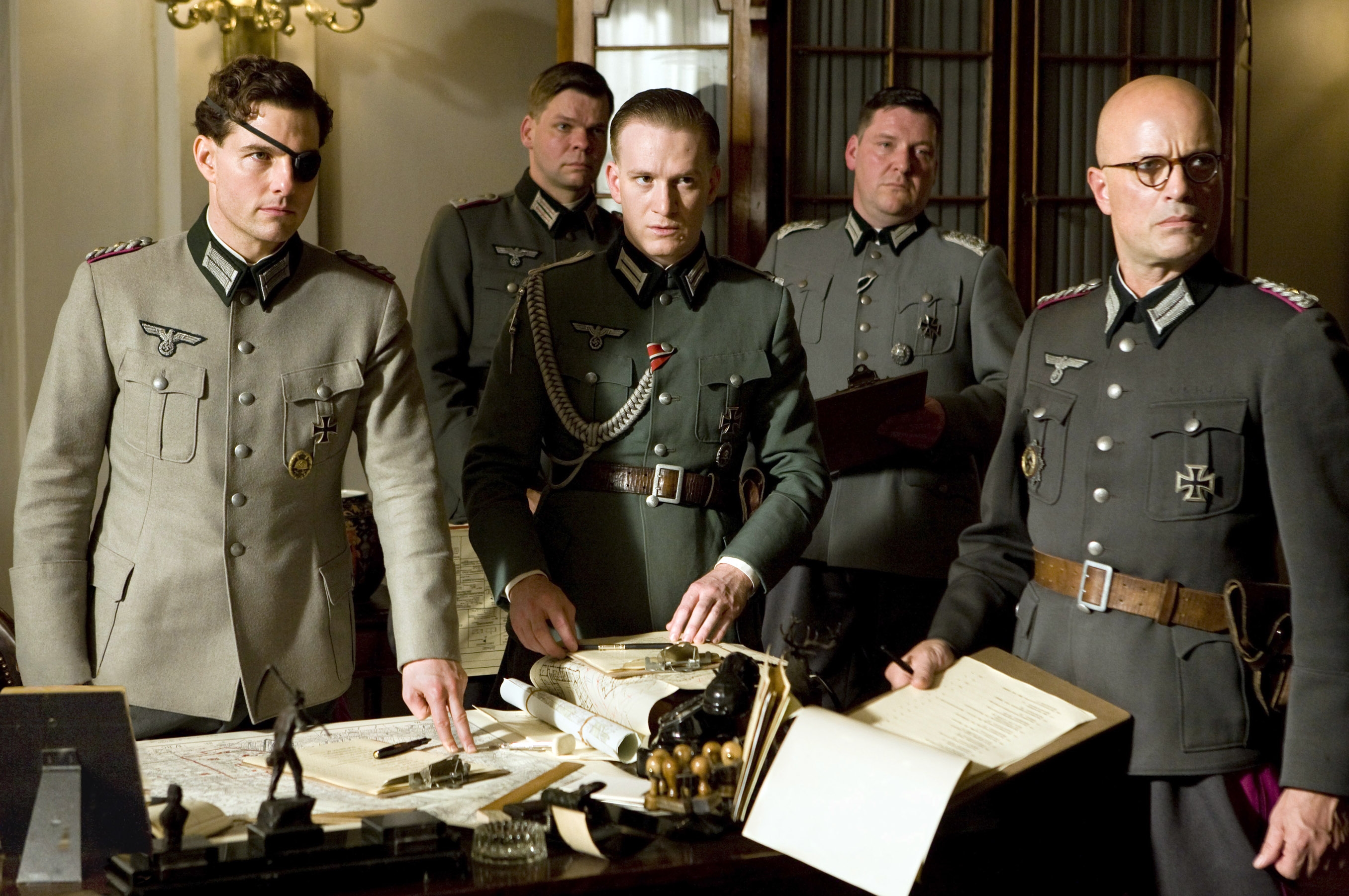 Five actors in WWII military uniforms gathered around a table with maps and documents, portraying a wartime strategy scene