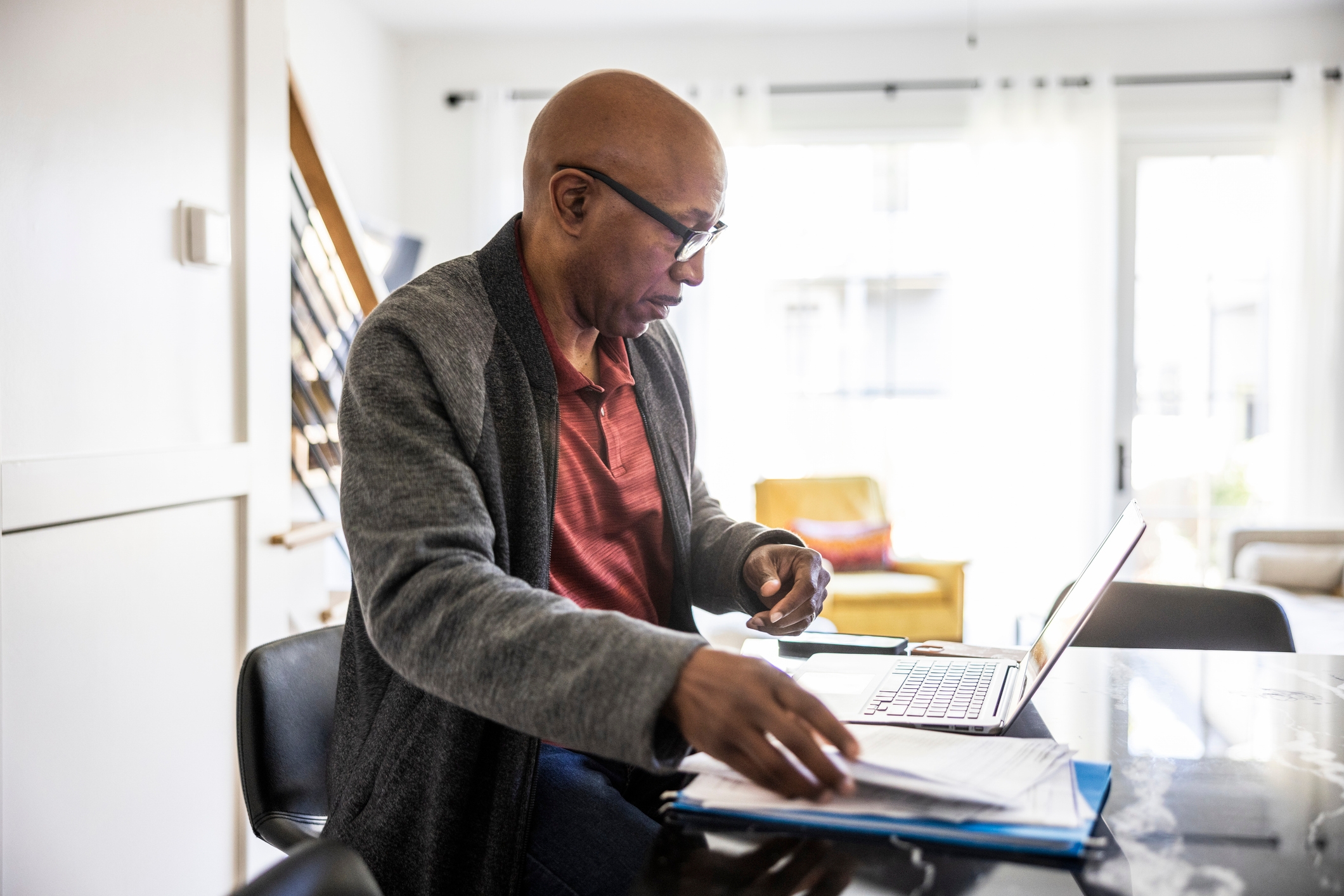 A person in casual attire sits at a desk, focused on a laptop and sorting through papers in a modern, well-lit home office