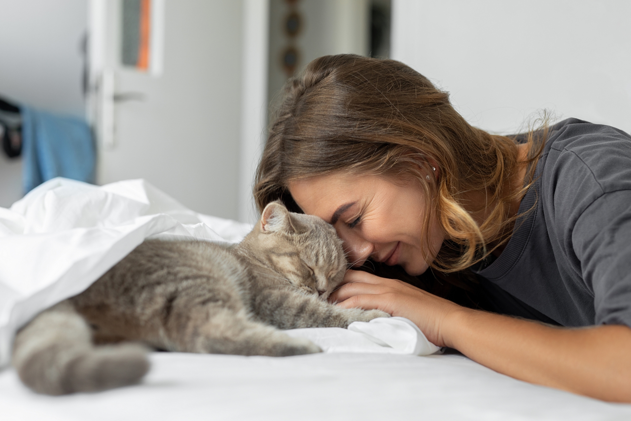 Woman gently rests her head against a sleeping cat on a bed, both appearing relaxed and content
