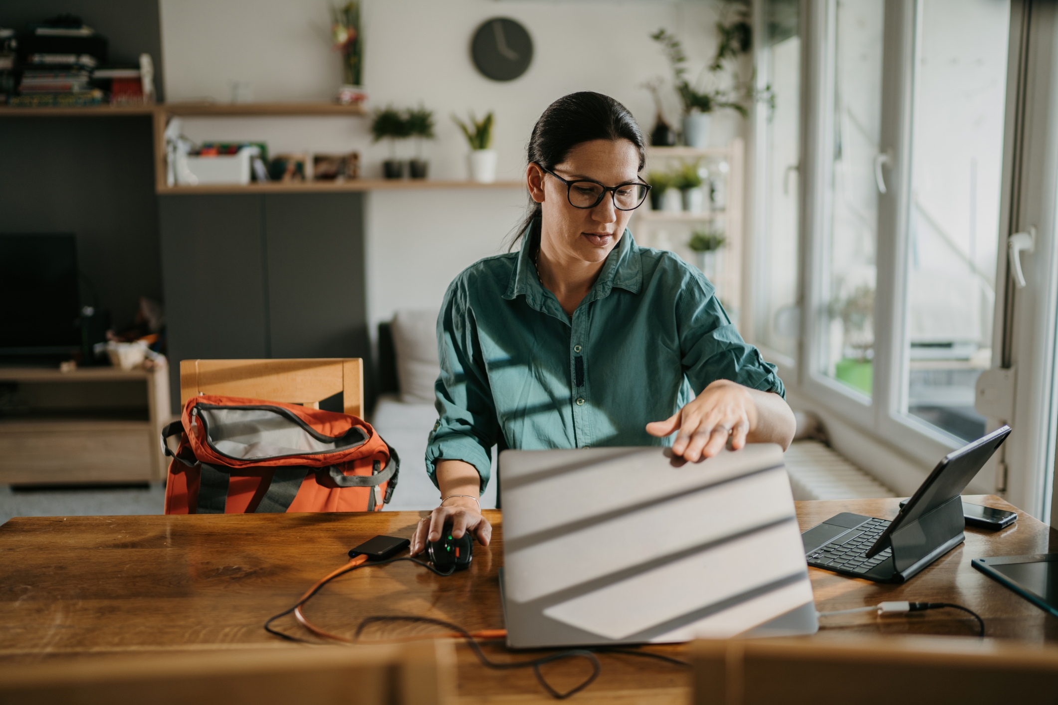 A person is working on a laptop at a wooden table, with a tablet, phone, and backpack nearby. Sunlight streams through the window