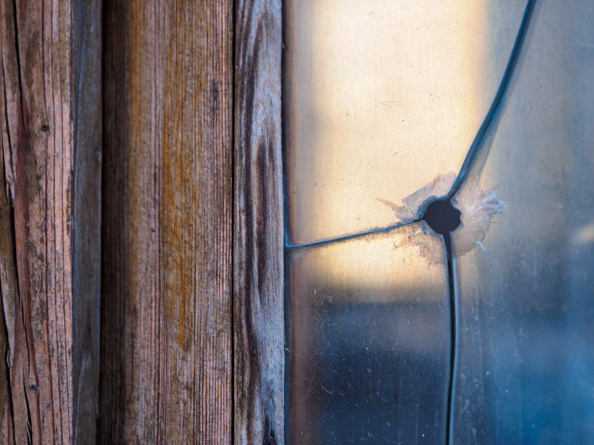 Cracked windowpane in a wooden frame with a circular hole, suggesting impact. Weathered wood texture contrasts with smooth glass surface