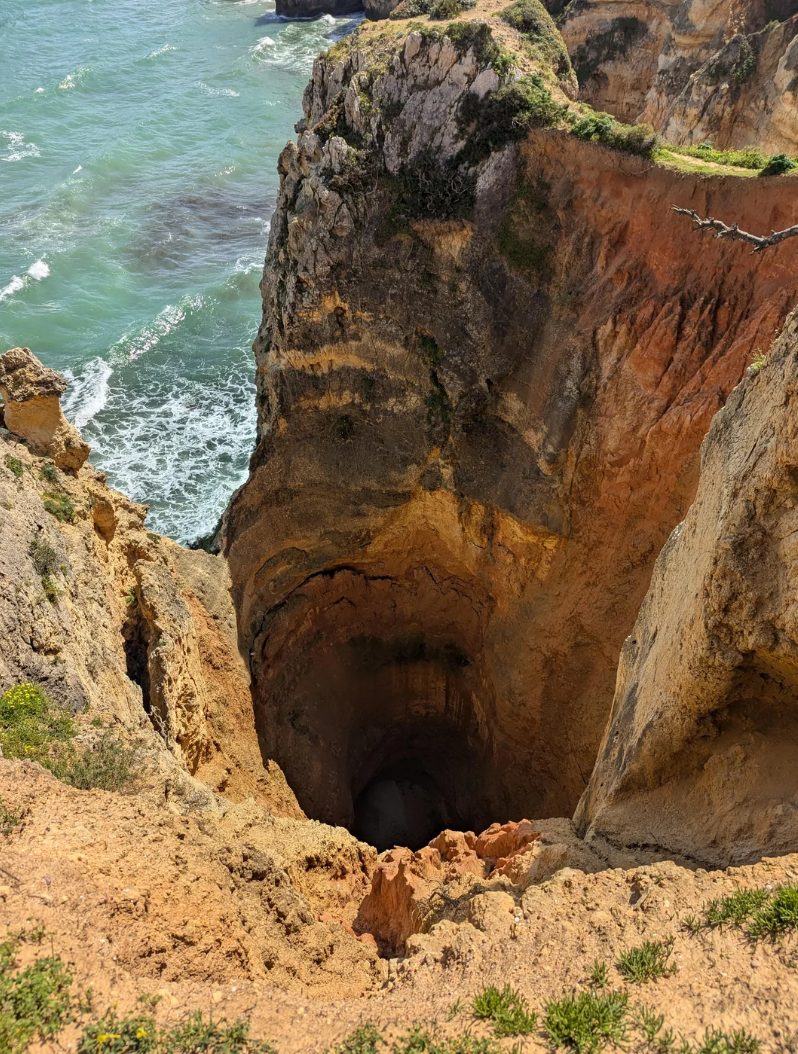 Steep coastal cliff with a deep, wide hole, overlooking ocean waves below. Grass and rough terrain surround the cliff edge