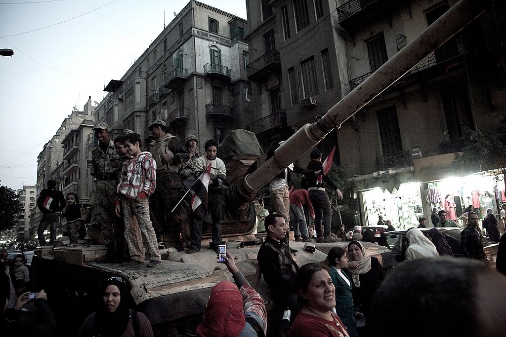 People gather on and around a military tank in an urban street, holding flags and taking photos, indicating a public demonstration or event